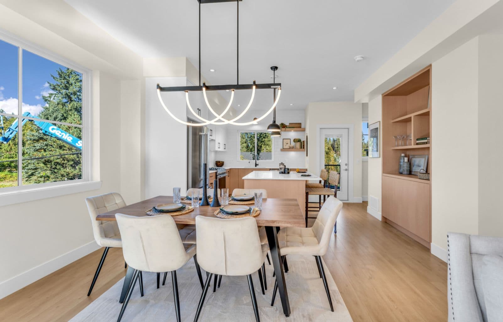 The Warwick – Open-concept dining and kitchen with a wood table set for a meal, arched pendant light fixture, white kitchen with island, and built-in wood shelving to the side.