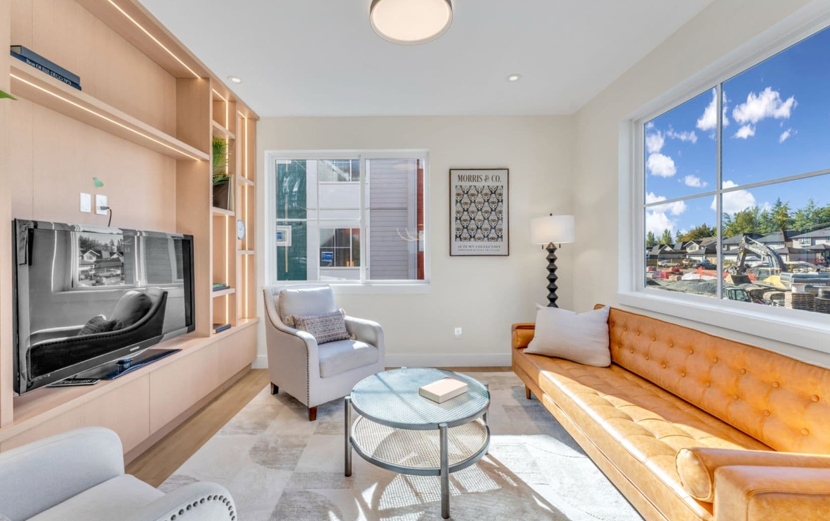The Warwick – Living room with a mustard leather tufted sofa, built-in wood media wall with TV, an armchair, and large windows looking out to a construction site.