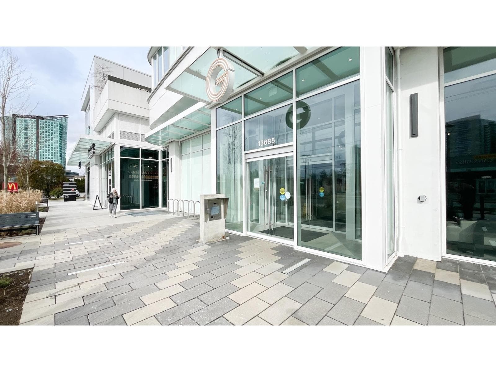 Georgetown One – A ground-level view of a modern residential building's entrance featuring double glass sliding doors, a glass canopy overhead, and prominent street address numbering. The surrounding area includes a wide, patterned stone paved walkway, stainless steel bike racks, and high-rise towers visible in the background under daylight.