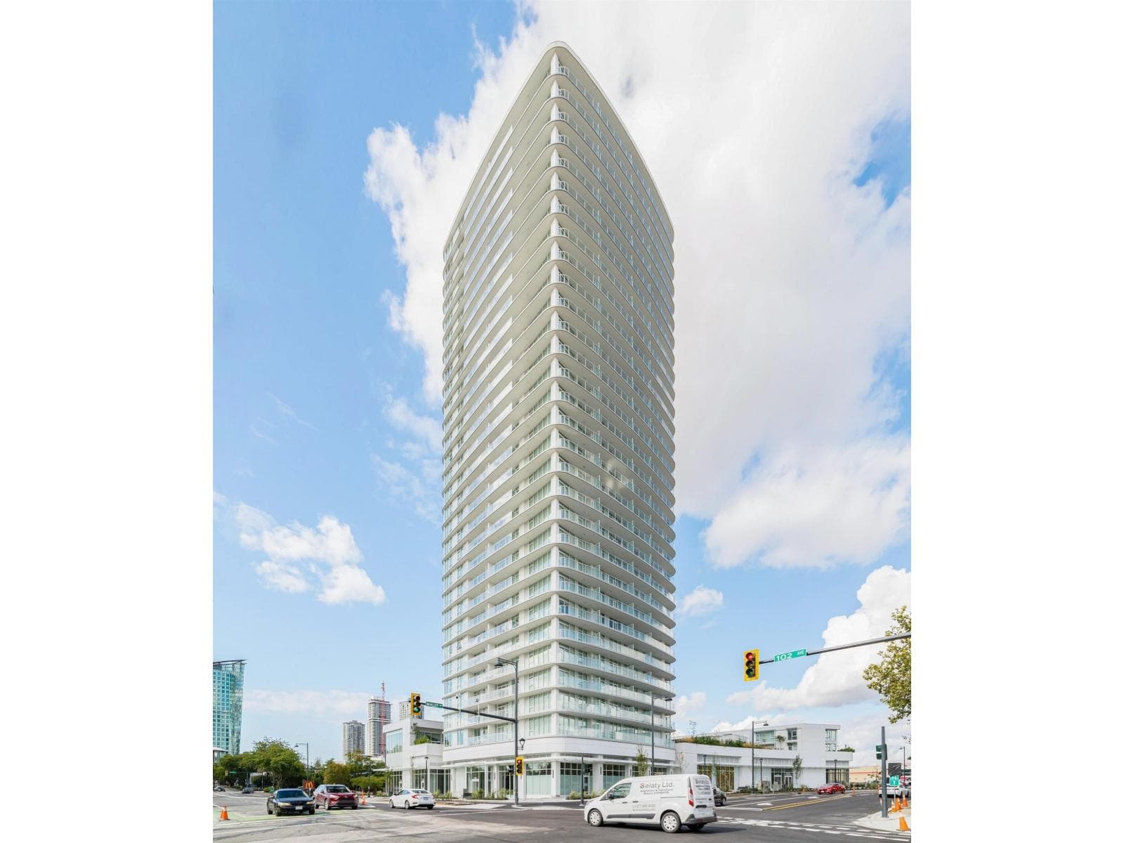 Georgetown One – A ground-level, low-angle shot of a sleek white high-rise residential tower with curved glass balconies standing at a city intersection under a bright blue, partly cloudy sky. Street-level retail spaces, traffic lights, cars, and surrounding city buildings are visible along the road in front of the structure.