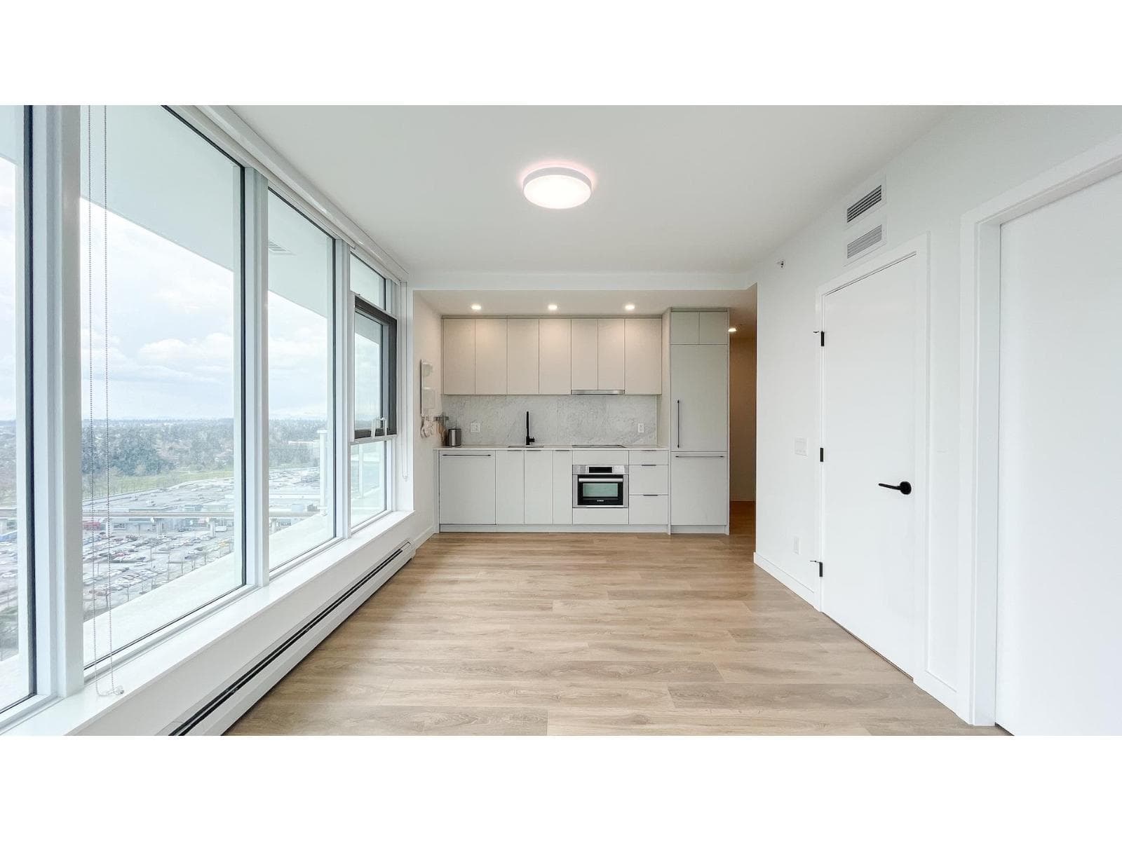 Georgetown One – An open-concept condo interior featuring light hardwood floors, crisp white walls, and a wall of floor-to-ceiling windows overlooking an urban vista. The rear kitchen is equipped with minimalist white cabinetry, integrated stainless steel appliances, a black faucet, and a stone backsplash.