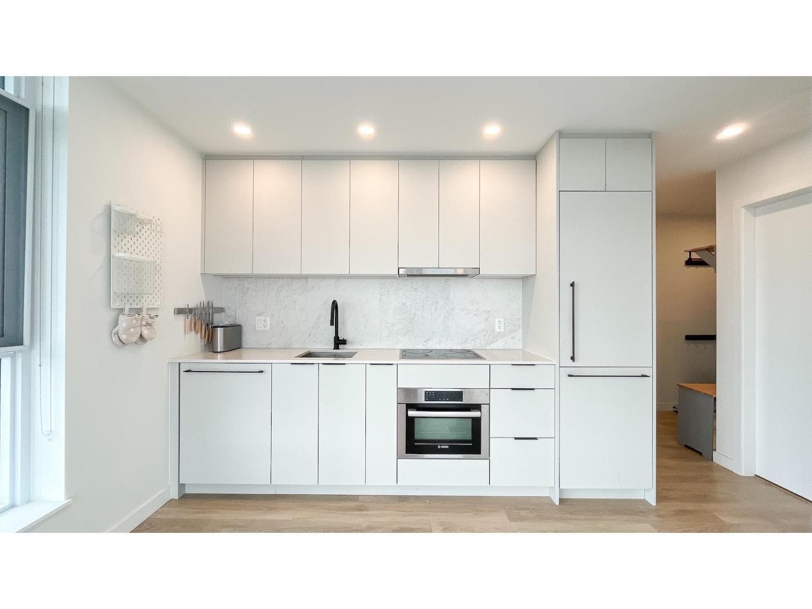 Georgetown One – A modern, minimalist kitchen features flat-panel white cabinetry, a integrated panelled refrigerator, and light wood flooring. The workspace includes a stainless steel oven, a grey ceramic cooktop, a matte black gooseneck faucet over an undermount sink, and a white marble backsplash.