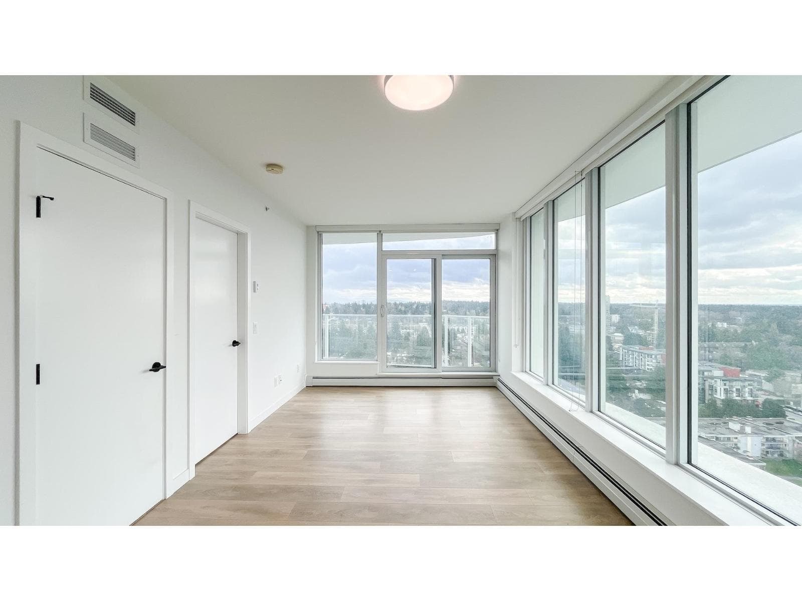 Georgetown One – An empty, bright condominium living area featuring light wood-grain laminate flooring, plain white walls, and two interior doors with black handles. Expansive floor-to-ceiling windows and glass sliding doors lead to a balcony, offering elevated views of a suburban landscape under an overcast sky.