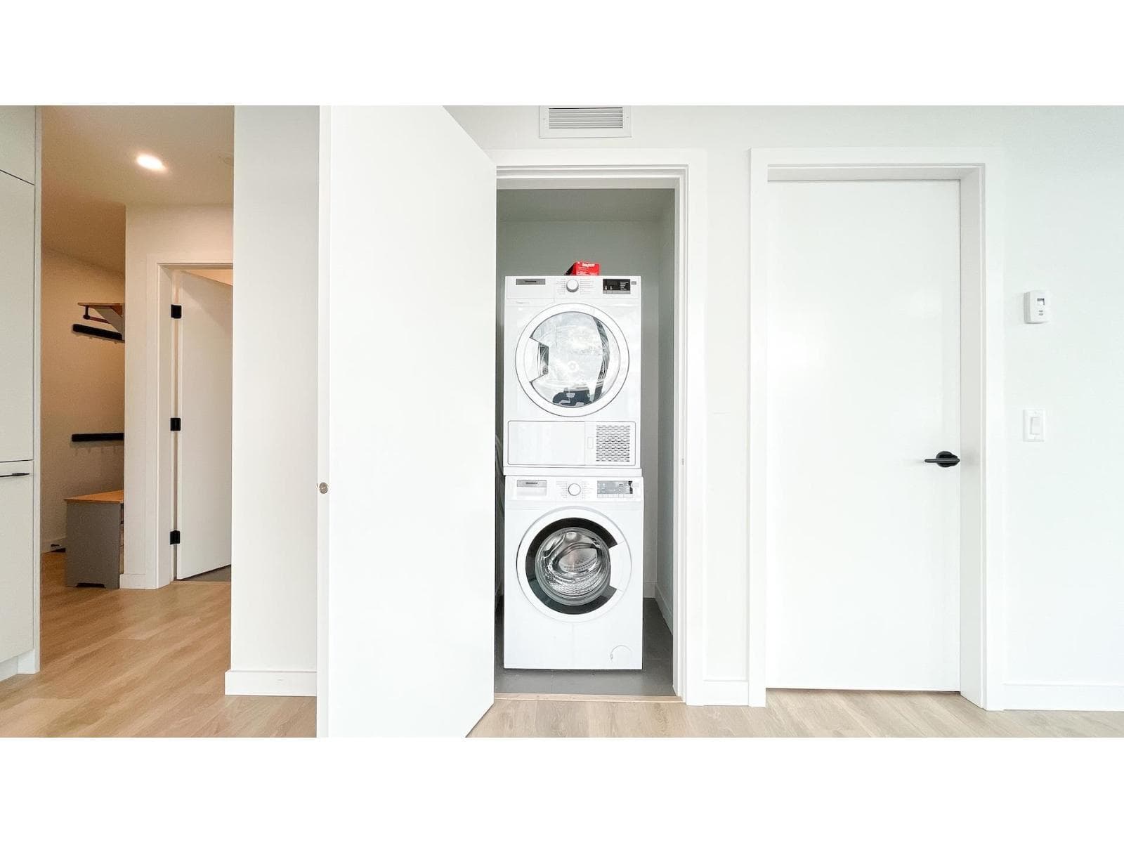 Georgetown One – A modern residential interior showing a stacked white washer and dryer neatly tucked inside a laundry closet with a wide open white door. Clean white walls and light hardwood flooring extend through the hallway, leading past another interior door toward an adjacent bedroom closet.