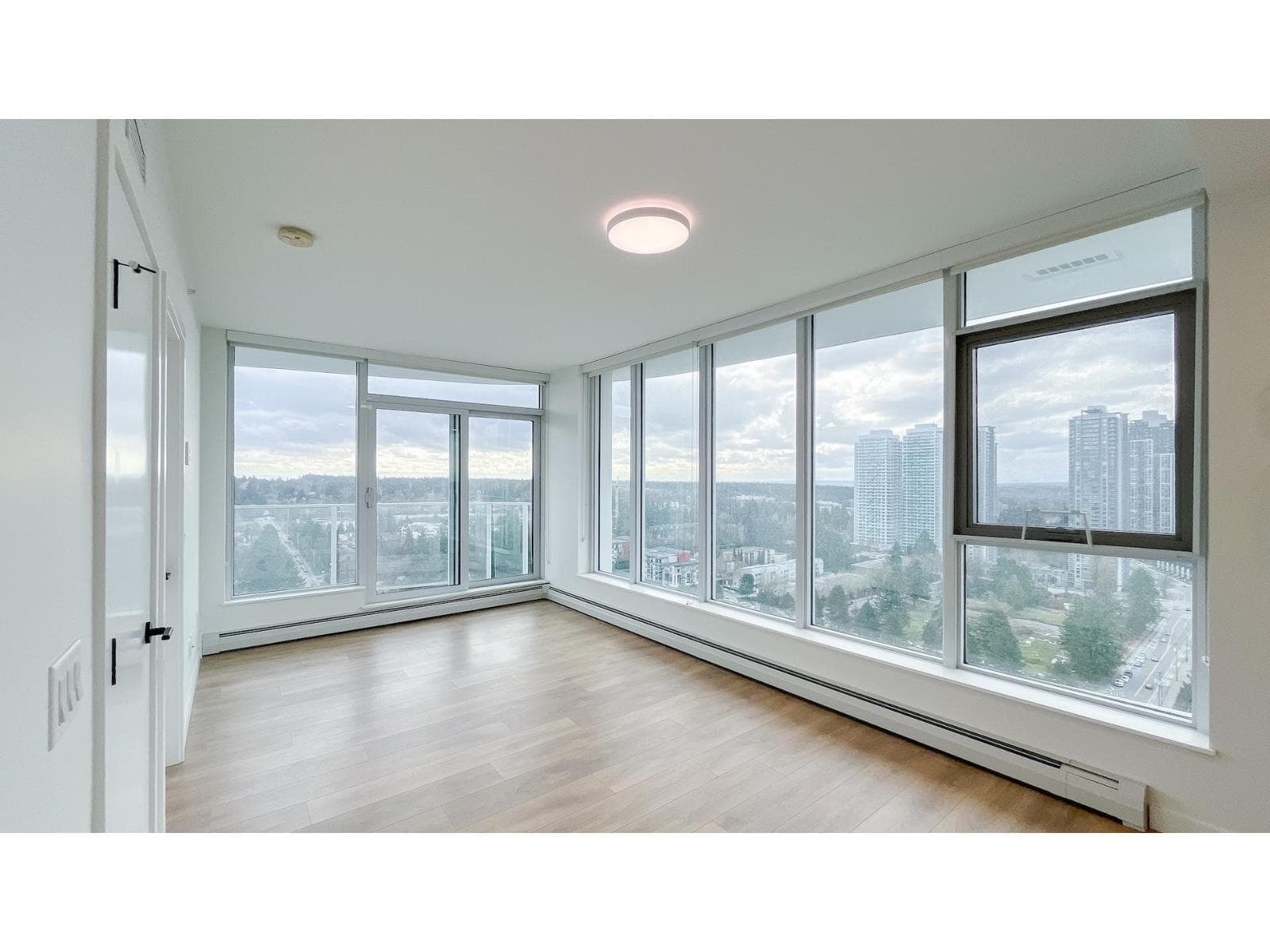 Georgetown One – An empty, bright condominium room featuring light wood-laminate flooring, white walls, and expansive floor-to-ceiling windows. The corner windows offer an elevated view of surrounding high-rise buildings and suburban greenery under an overcast sky.