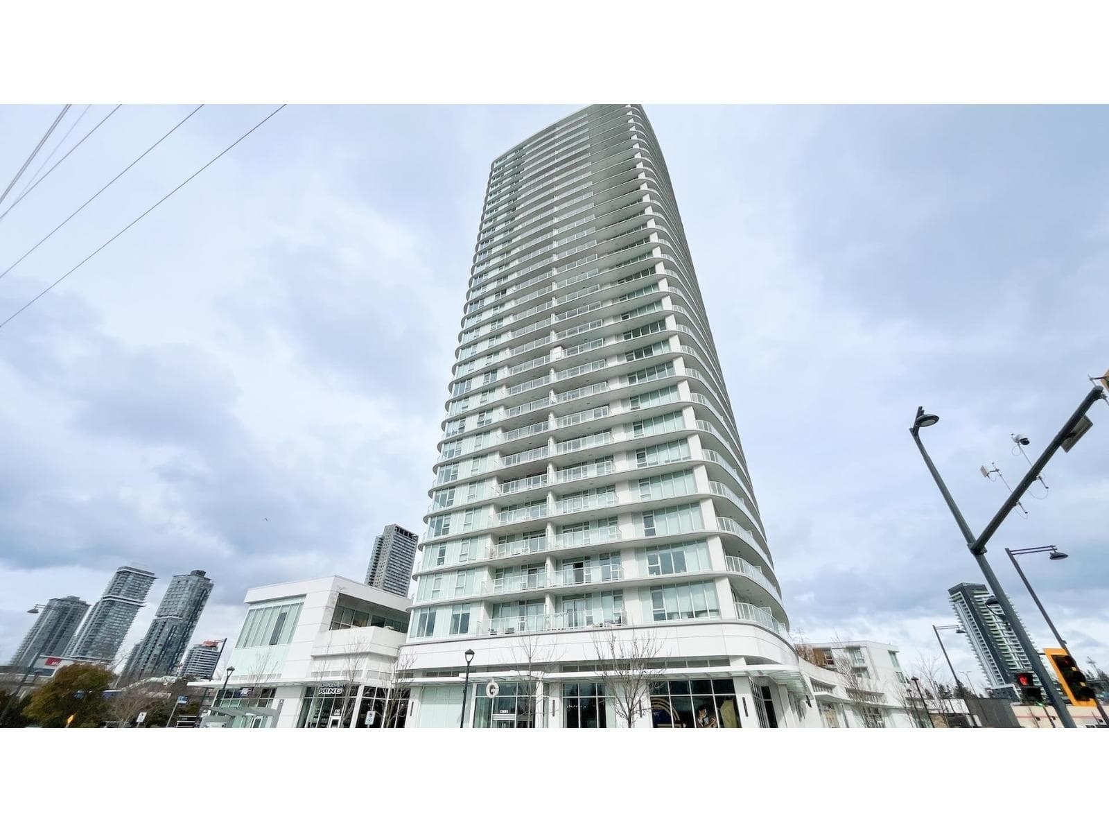 Georgetown One – A low-angle shot showcases a tall, modern residential tower with curved white glass balconies extending upward against a cloudy sky. The ground level features retail spaces with large glass storefronts along the street, set in an urban environment with neighboring high-rises in the distance.
