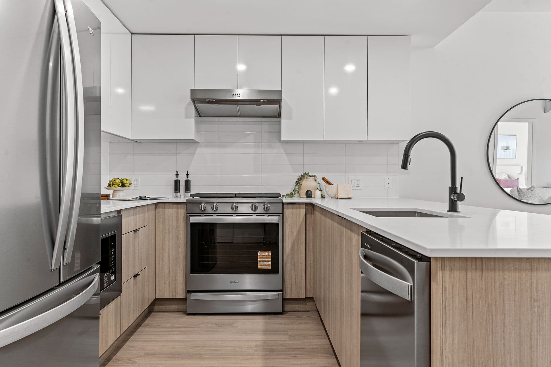 Hawthorne – A modern U-shaped kitchen featuring light wood lower cabinets, high-gloss white upper cabinets, and white quartz countertops with a matching tile backsplash. The space is equipped with stainless steel appliances, a matte black gooseneck faucet over an under-mount sink, and subtle decorative accents on the counter.