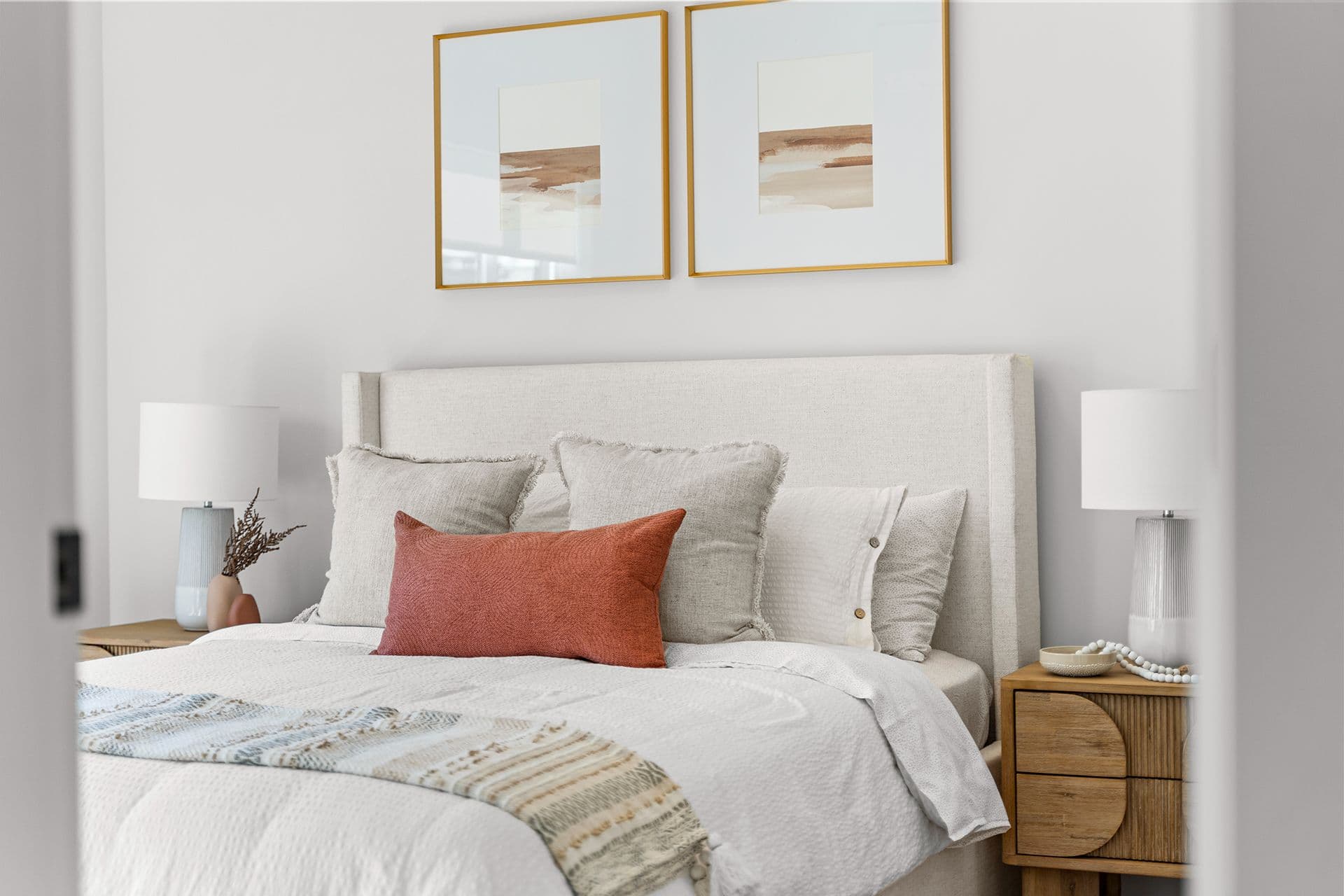 Hawthorne – A bright, tastefully styled bedroom features a bed dressed in neutral white linens, fringe-trimmed accent pillows, and a rust-orange lumbar cushion against a fabric upholstered headboard. Flanking wooden nightstands with ribbed drawers hold sleek table lamps, while two framed abstract art prints hang symmetrically on the light grey wall above.