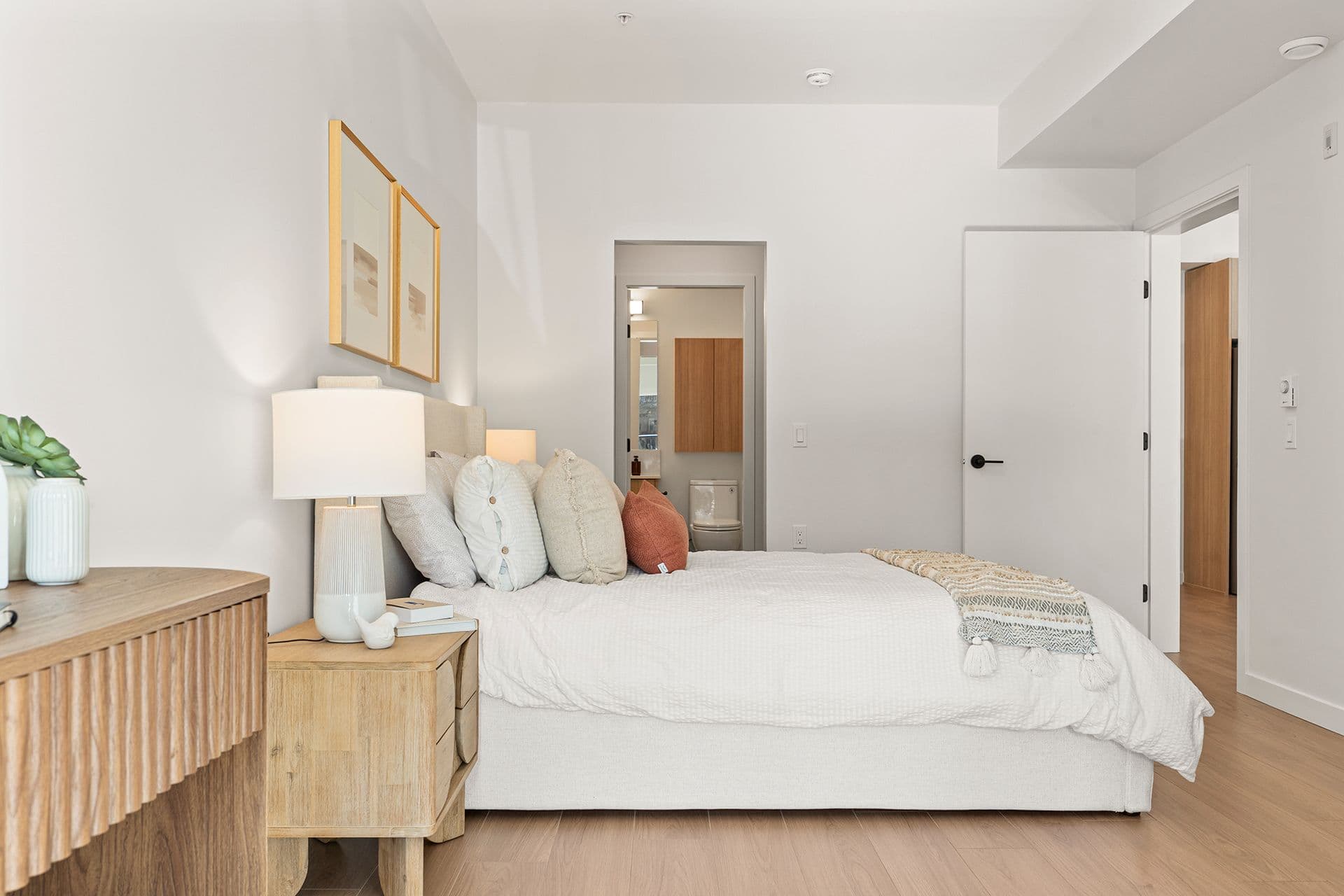 Hawthorne – A bright, modern bedroom featuring a neatly made bed with light-colored linens, accent pillows, and wooden nightstands with soft lamp lighting. The room is decorated in neutral tones with light oak flooring and offers a doorway view into an ensuite bathroom.