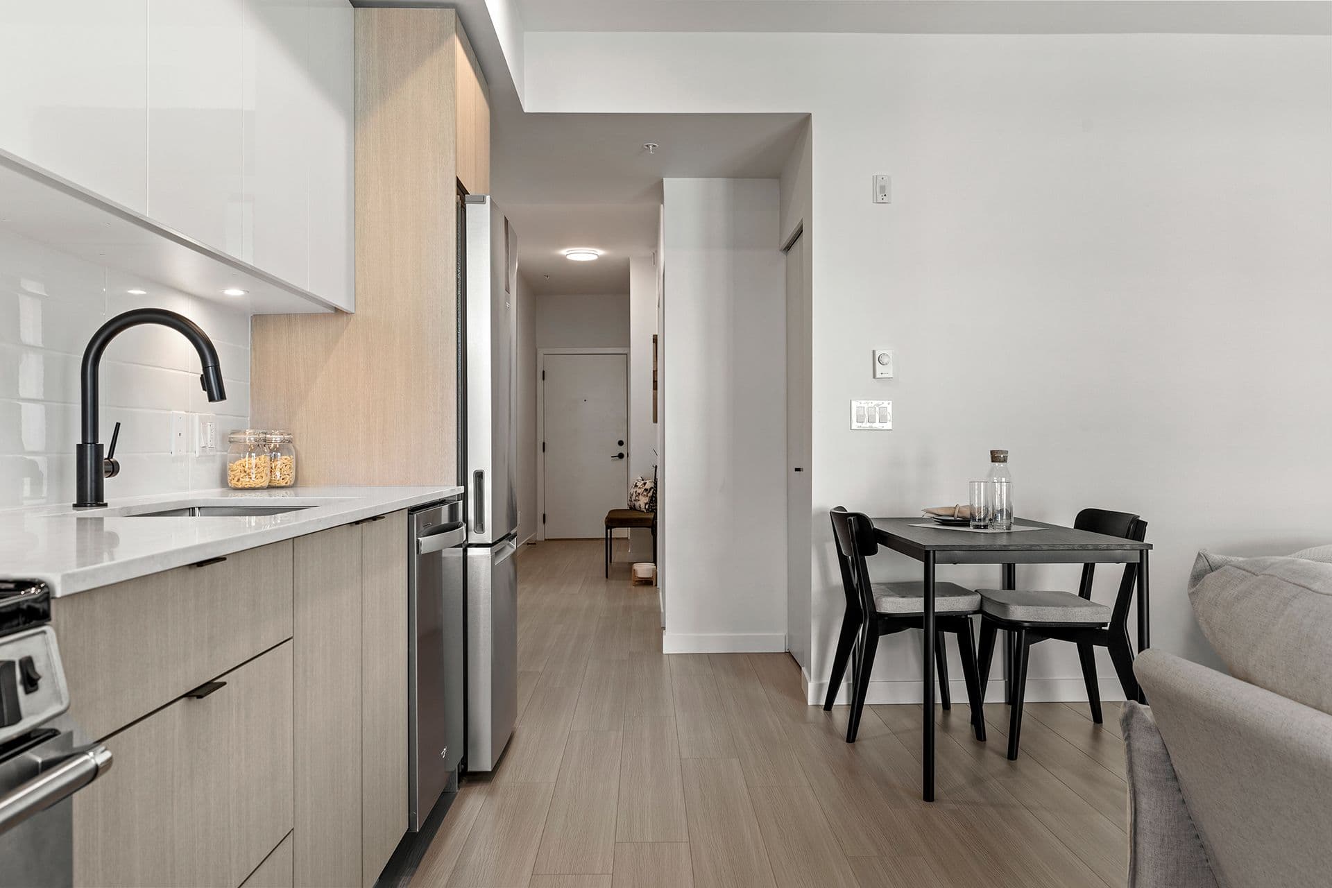 Hawthorne – A modern condominium interior looking past a sleek kitchen toward a small dining nook and hallway entrance. The kitchen features flat-panel light wood and glossy white cabinetry, stainless steel appliances, white quartz countertops, and a matte black faucet, while light laminate flooring seamlessly connects the dining table and living area.