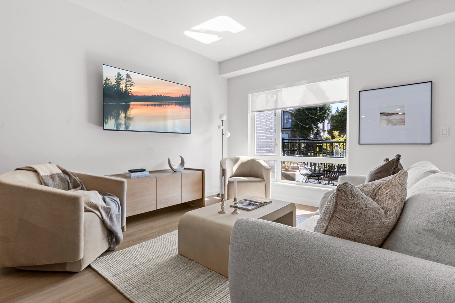 Hawthorne – A modern living room furnished in neutral tones featuring a light gray textured sofa, plush cream armchairs, a low coffee table, and a wall-mounted flat-screen TV above a light wood console. Natural light fills the space through a large window that looks out onto a patio and residential neighborhood.