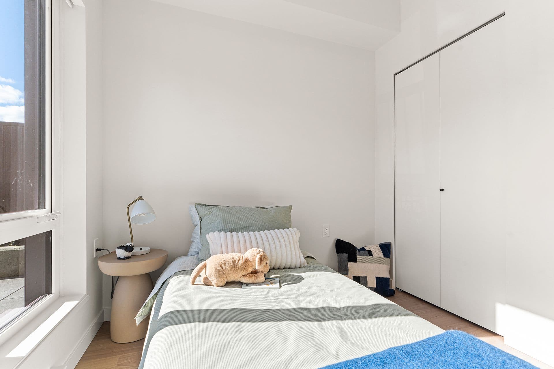 Hawthorne – A bright, compact bedroom feature a single bed dressed in sage green bedding, decorative pillows, and a stuffed animal. Sunlight streams through a large side window onto the bed, illuminating white walls, a modern round nightstand with a lamp, and sleek white closet doors.