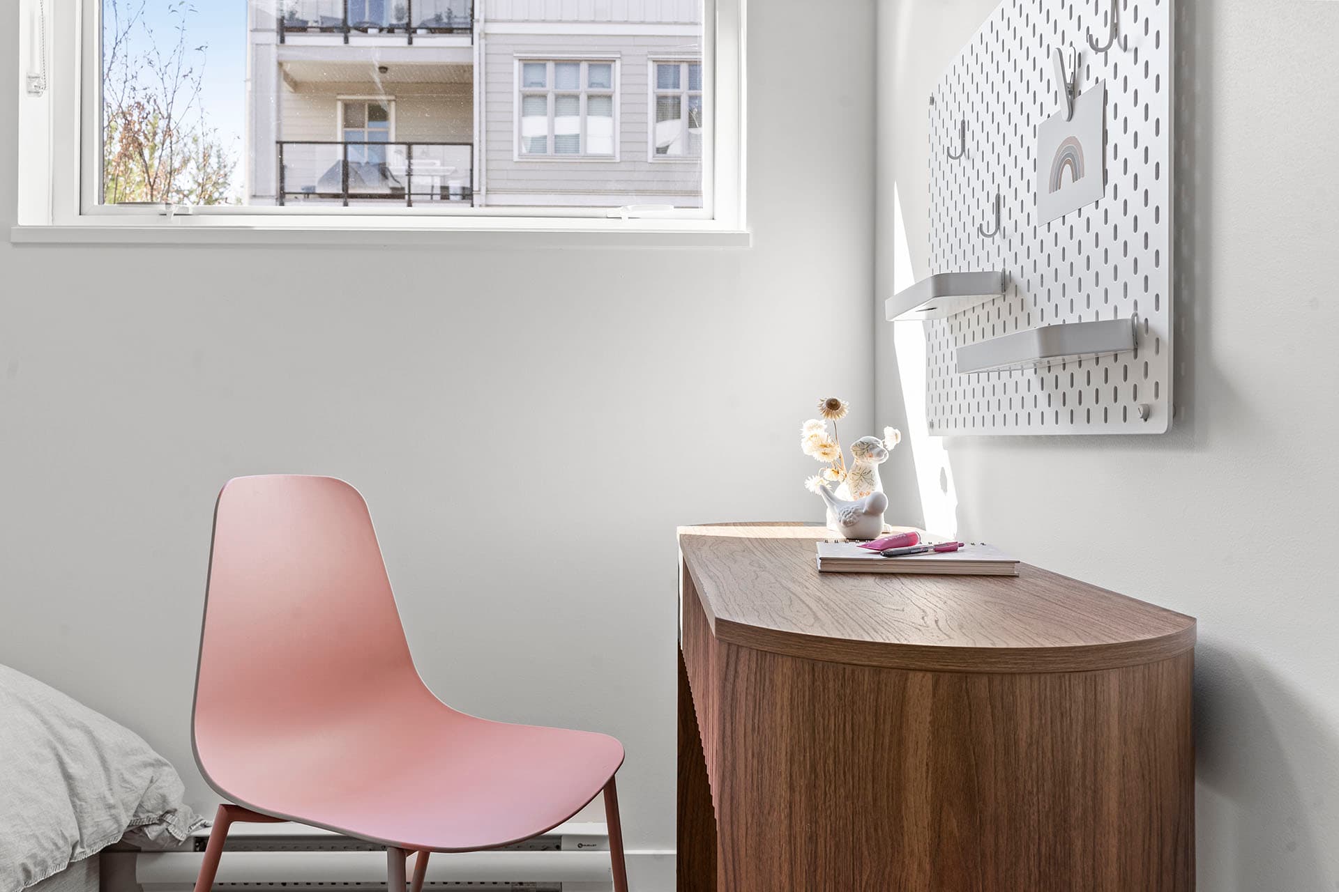Hawthorne – A bright home office corner features a curved wooden desk paired with a dusty pink accent chair next to a window filled with natural sunlight. On the white wall above the desk hangs a metal pegboard organizer with small shelves, hooks, and stationery accessories.