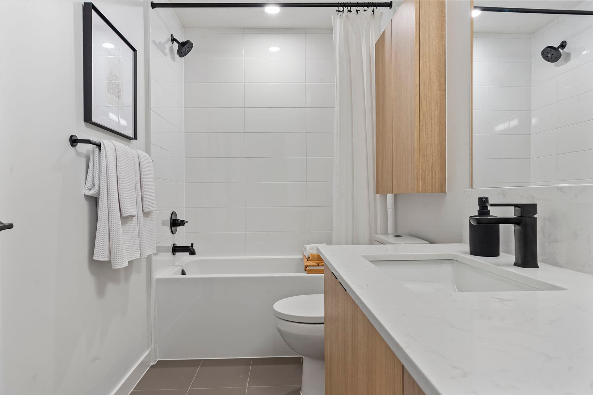 Hawthorne – A modern residential bathroom featuring a white bathtub with subway-tiled walls, matte black fixtures, and a light wood vanity topped with a white marble quartz countertop. Bright receded lighting and neutral tile flooring create a clean, minimalist, and serene atmosphere.