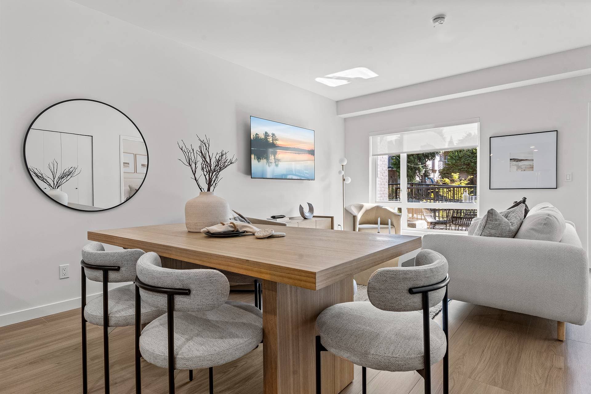 Hawthorne – A modern open-concept dining and living room area features a wooden dining table paired with stylish grey fabric chairs set against light wood-look flooring and white walls. In the background, a light grey sofa sits near a large window overlooking an outdoor patio surrounded by green trees.
