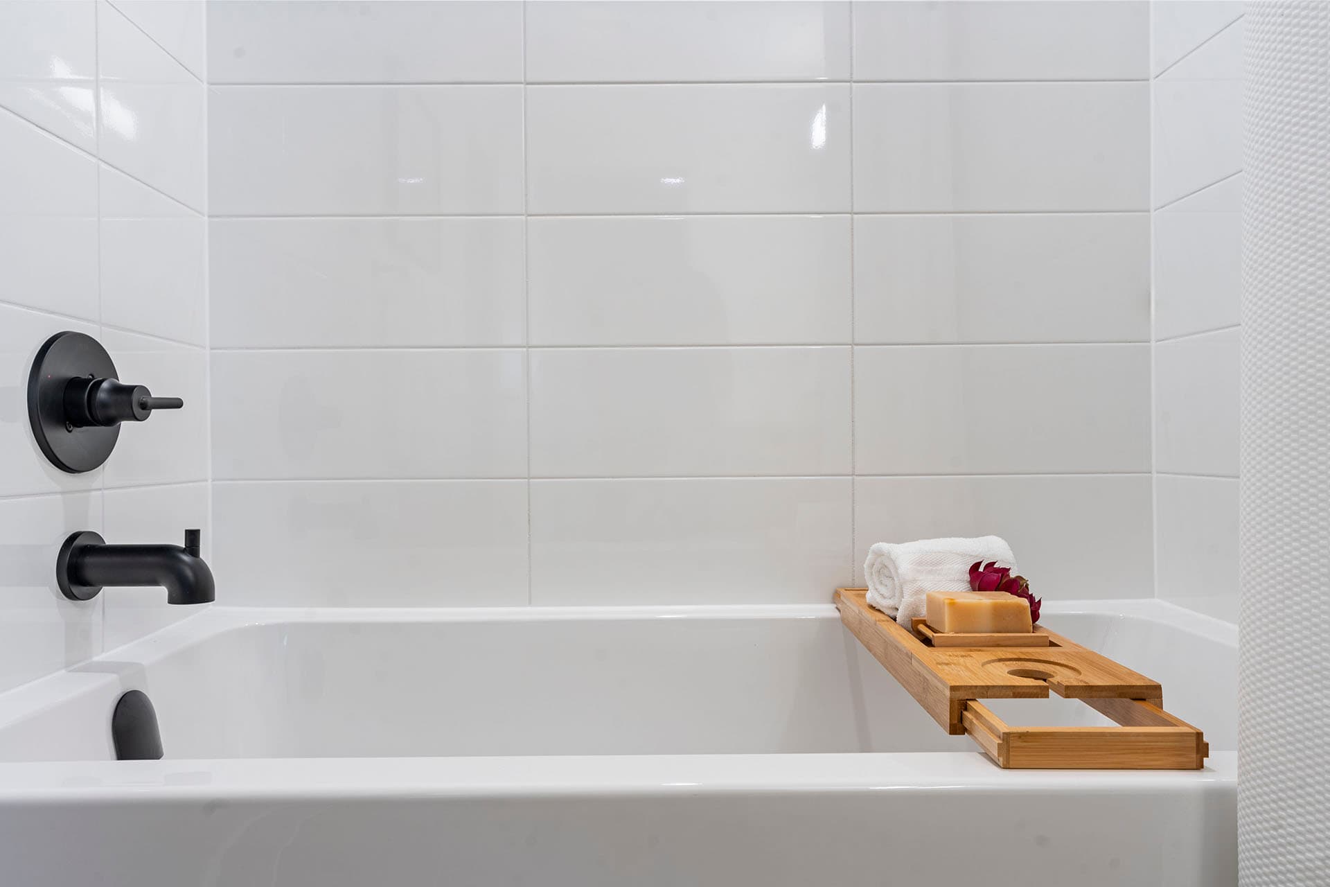 Hawthorne – A clean, modern bathtub setup featuring glossy white subway wall tiles and matte black plumbing fixtures. A wooden bath caddy rests across the tub, holds a rolled white towel, bar soap, and small decorative flower, creating a bright, spa-like atmosphere.