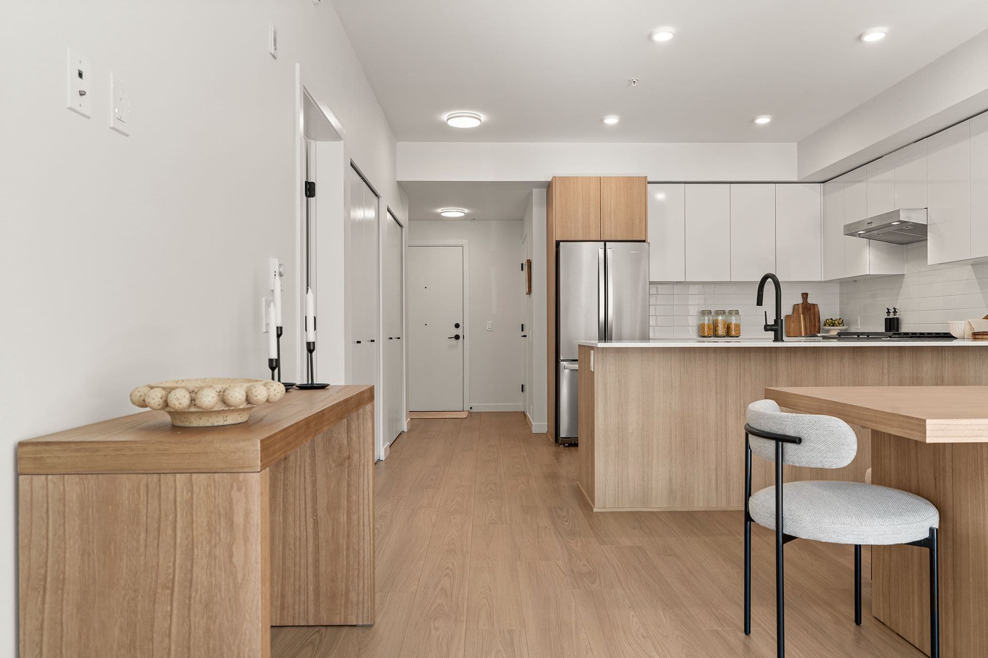 Hawthorne – A modern open-concept condo entryway and kitchen feature light wood-grain flooring, white walls, and flat-panel cabinetry in a mix of white and light wood tones. The kitchen includes stainless steel appliances, a white tile backsplash, a contrast black faucet, and a large island facing an adjacent dining area.
