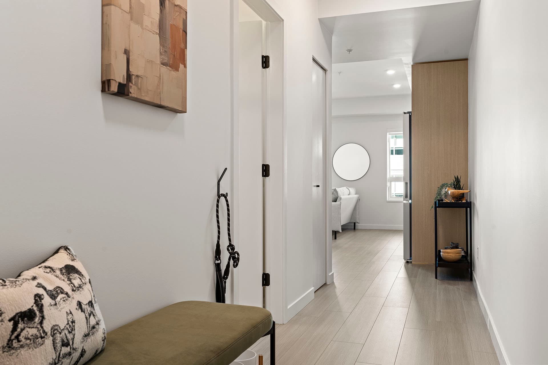 Hawthorne – A bright, narrow residential hallway with light wood-look flooring and white walls leads from an entryway bench toward an open living space. Abstract wall art, a small decorative side table, and modern interior doors create a clean, minimalist contemporary feel.