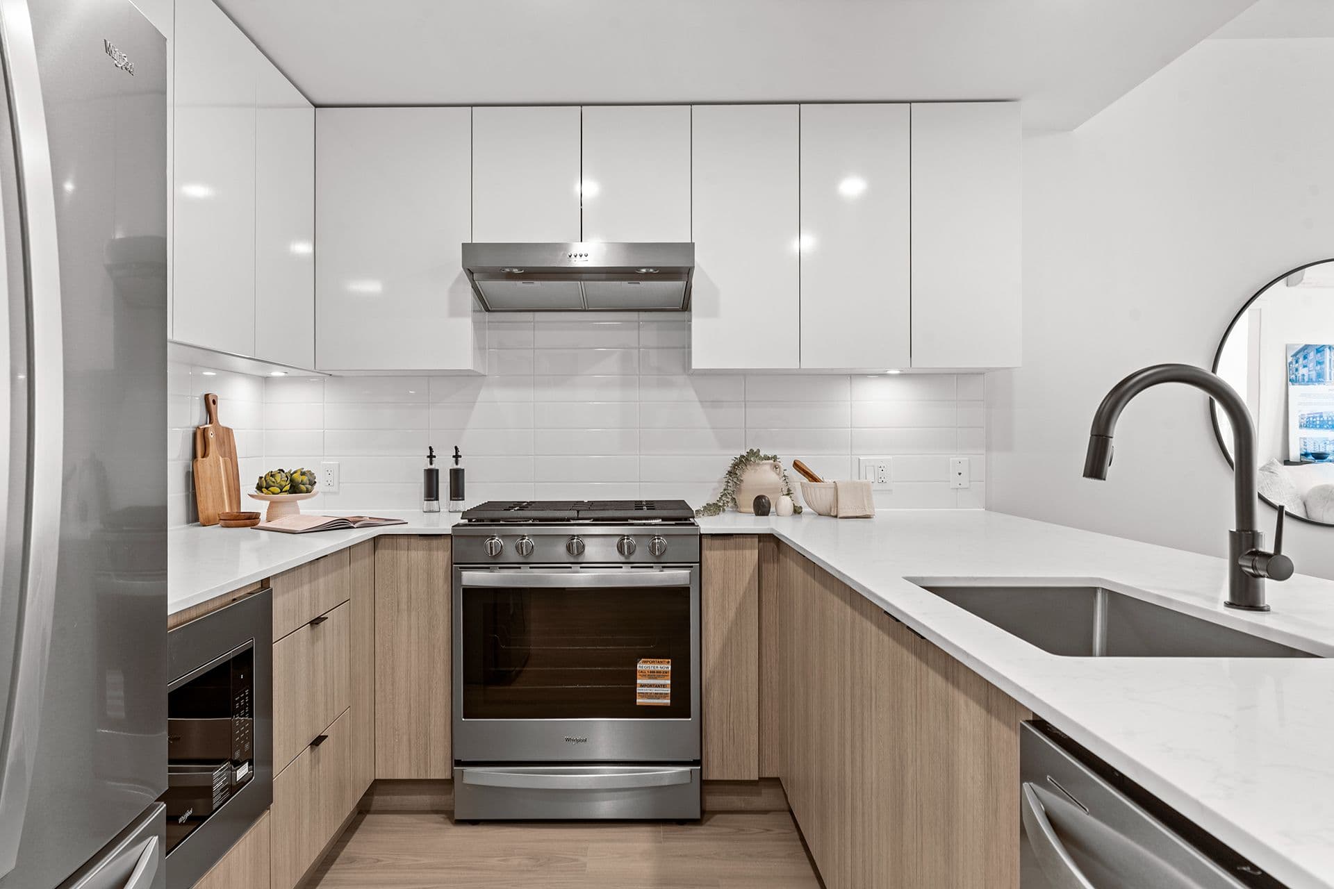 Hawthorne – A modern U-shaped kitchen features glossy white upper cabinets and light wood lower cabinetry paired with white quartz countertops and a white tile backsplash. Stainless steel appliances include a gas range, range hood, built-in microwave, and a black gooseneck faucet over an undermount sink.