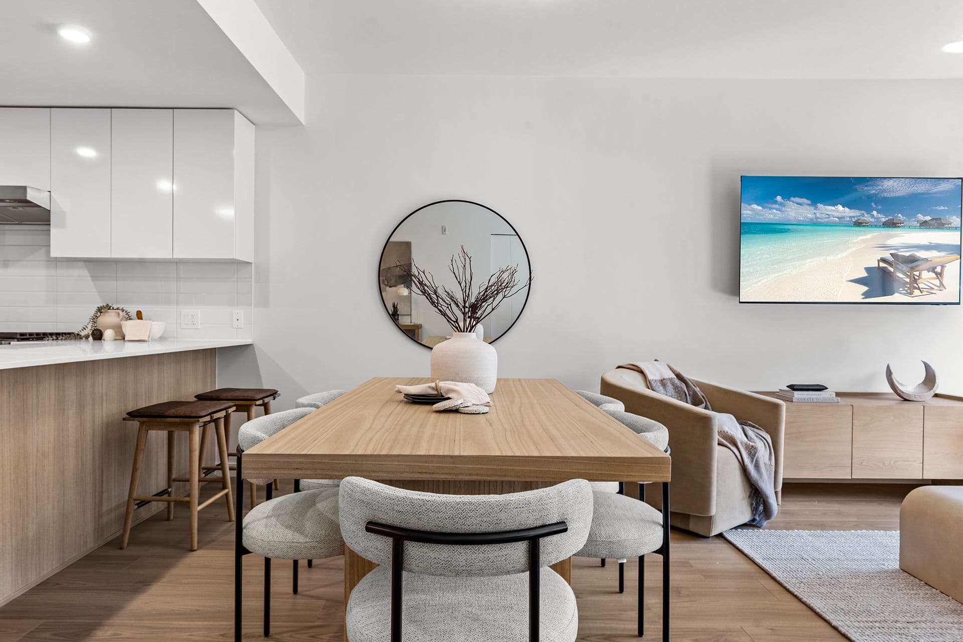 Hawthorne – A modern open-concept dining and living space features a natural wood dining table surrounded by upholstered gray chairs, with a kitchen island and barstools to the left. Bright, neutral tones dominate the room, highlighted by a wall-mounted round mirror, a flat-screen TV displaying a beach landscape, and smooth light-wood flooring throughout.