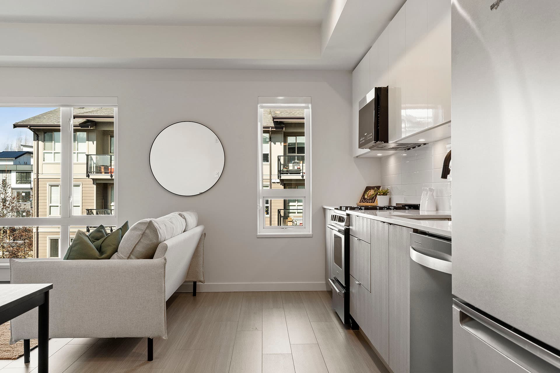 Hawthorne – A modern open-concept kitchen and living area featuring sleek grey flat-panel lower cabinets, glossy white upper cabinets, stainless steel appliances, and light laminate flooring. Natural daylight streams through large windows that look out onto neighboring residential buildings, illuminating a beige sofa with accent pillows and a circular wall mirror.