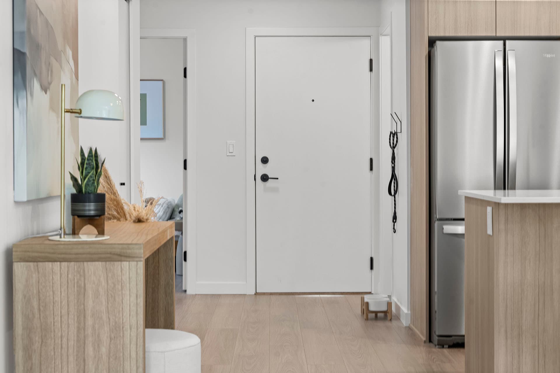 Hawthorne – A bright entry foyer featuring light wood flooring, white walls, and a plain white front door with black hardware. On the left is a wooden console table decorated with a table lamp and potted plant, while the right side opens to a kitchen area with light oak cabinetry and stainless steel appliances.