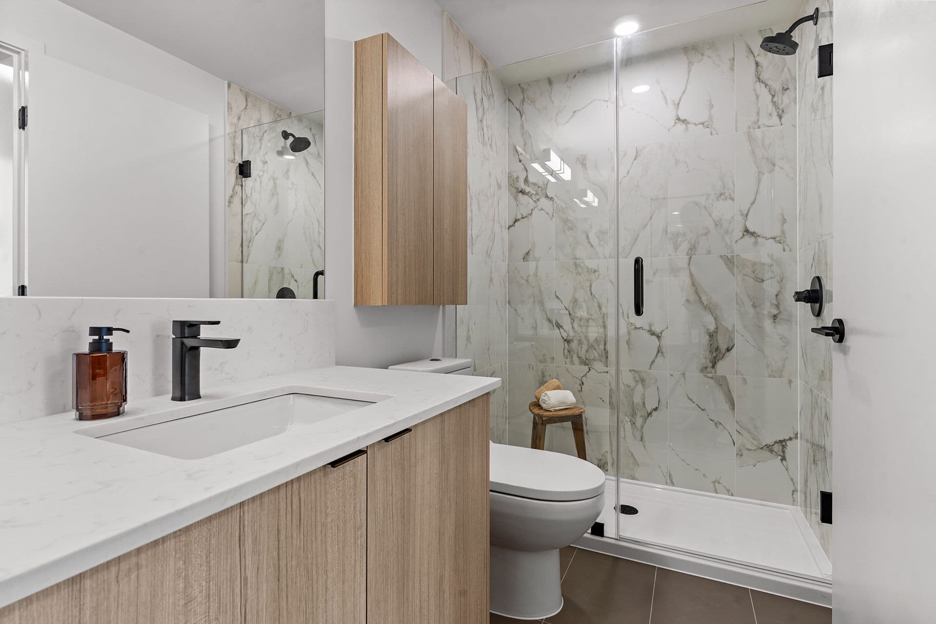 Hawthorne – A modern bathroom features light-toned wood cabinetry, matte black fixtures, and a white quartz vanity countertop with an undermount sink. A walk-in shower with frameless glass doors is lined with large white marble-patterned tiles, creating a clean and bright aesthetic.