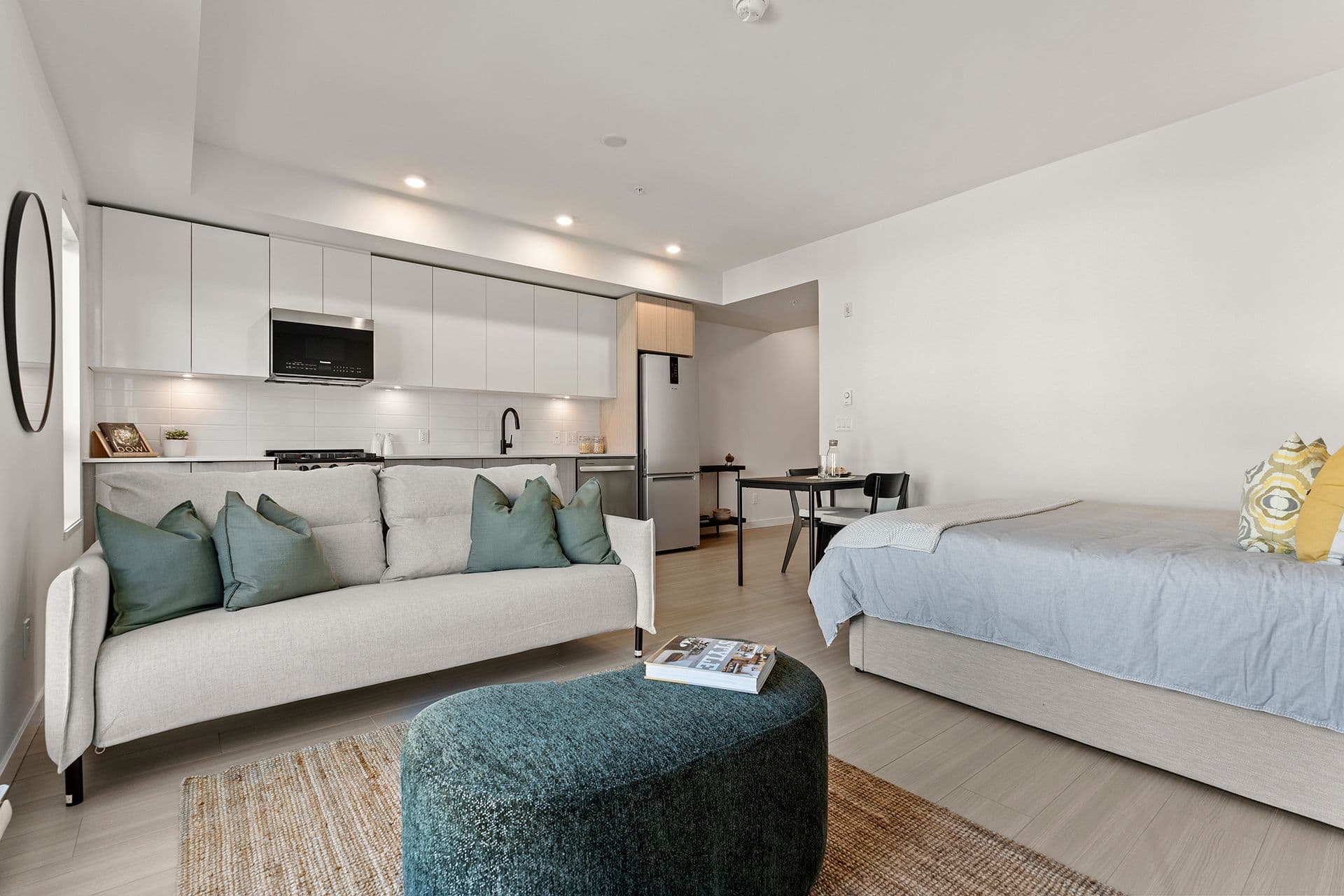 Hawthorne – A modern open-concept studio apartment featuring a light beige sofa with teal throw pillows, a textured oval ottoman, and a made bed with soft blue bedding. In the background, a linear kitchen with sleek white cabinetry, under-cabinet lighting, stainless steel appliances, and a small dining nook create a bright, contemporary living space.
