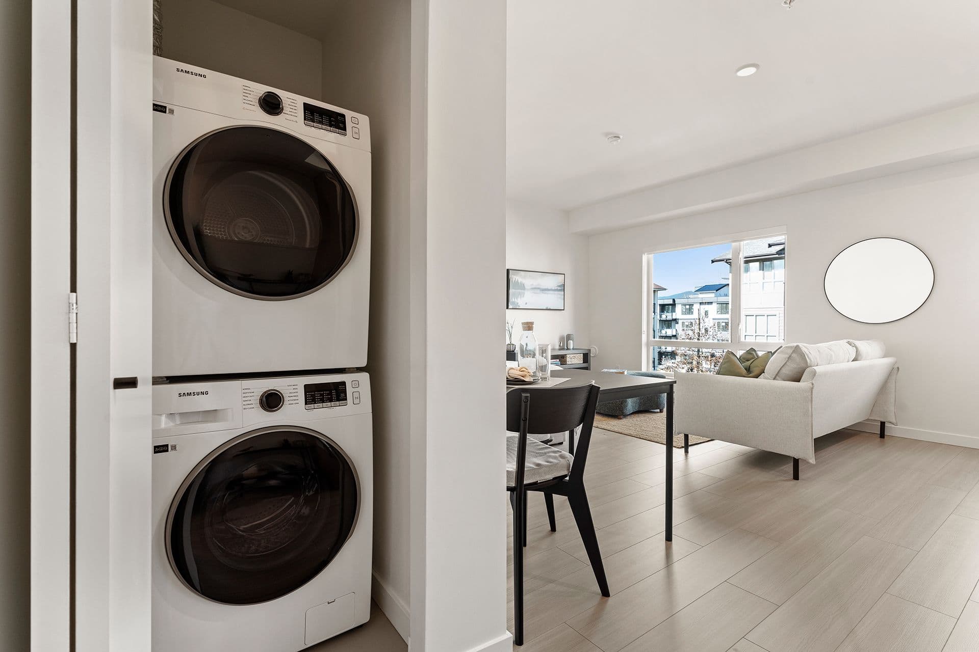 Hawthorne – A modern suite interior featuring stacked white Samsung front-loading washer and dryer units neatly tucked into an entry closet. Past the laundry nook, the open-concept layout reveals a dining set and a bright living area with light wood-look flooring, a beige sofa, and a large window looking out onto neighbouring buildings.