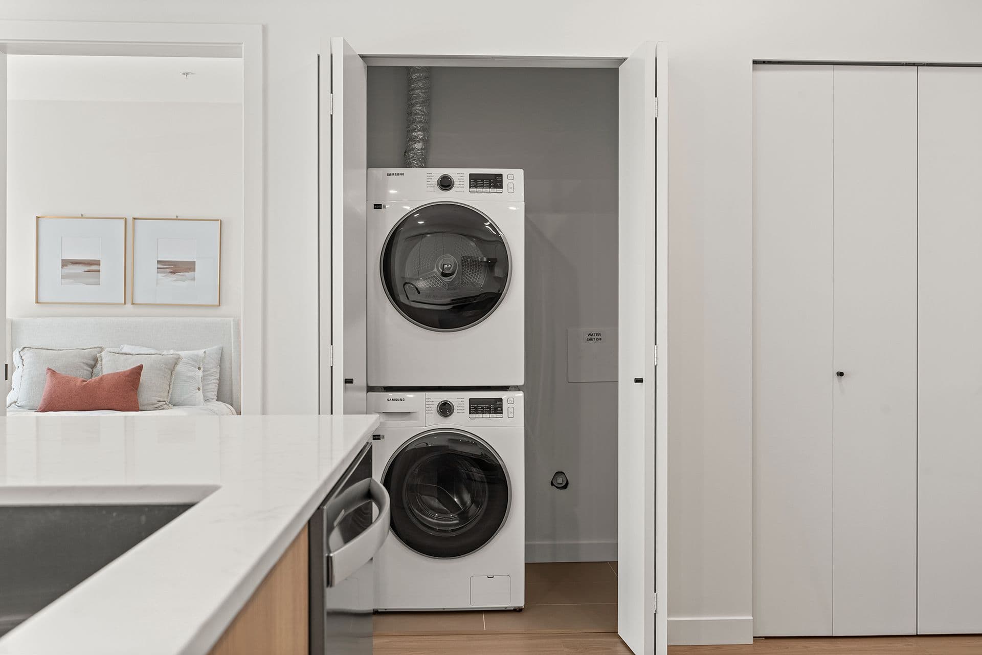 Hawthorne – A modern suite's bright white laundry closet featuring a stacked white Samsung washer and dryer set behind open bi-fold doors. The foreground shows an edge of a kitchen island countertop, while a doorway on the left provides a glimpse into a staged bedroom with framed art.