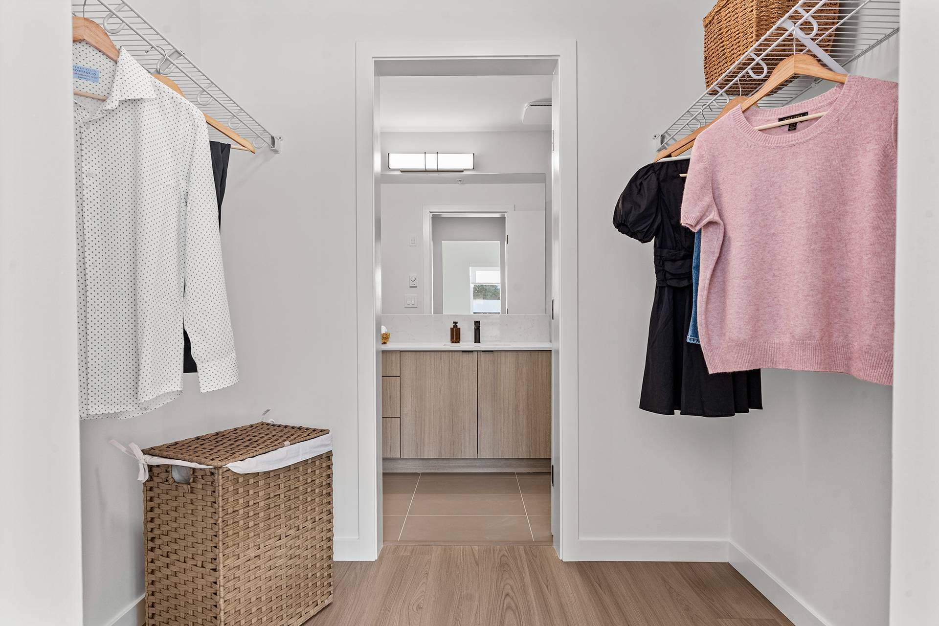 Hawthorne – A bright walk-in closet featuring wire organizer shelving with hung clothing, a woven hamper, and wood-look laminate flooring. Through the open doorway, an ensuite bathroom is visible with modern wood-grain cabinetry, light tile floors, and a lit mirror vanity.