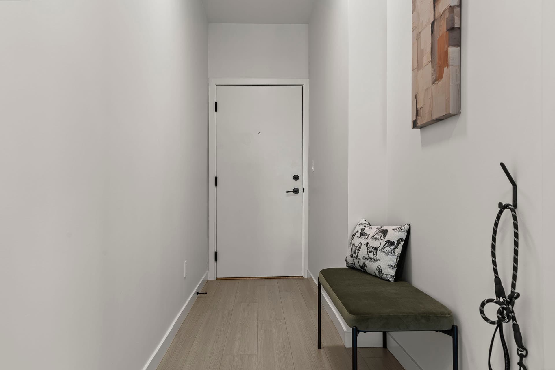 Hawthorne – A narrow residential entryway featuring white walls, light wood flooring, and a white front door with black hardware. A olive green padded bench with an accent pillow sits along the right wall below framed canvas art, with a black dog leash hanging nearby.