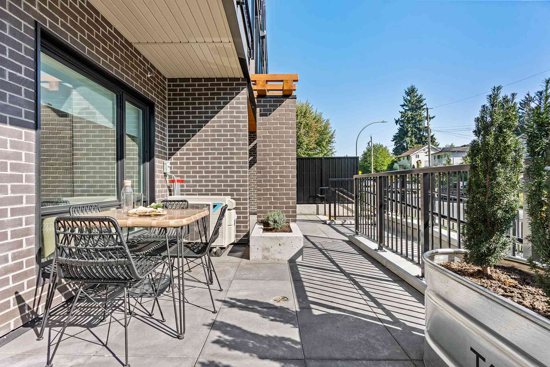Hawthorne – A ground-floor private patio features dark brick exterior walls, stone paver flooring, and a small woven outdoor dining set under a covered overhang. Metal railings and planter boxes border the space, looking out toward a sunny residential street lined with trees.