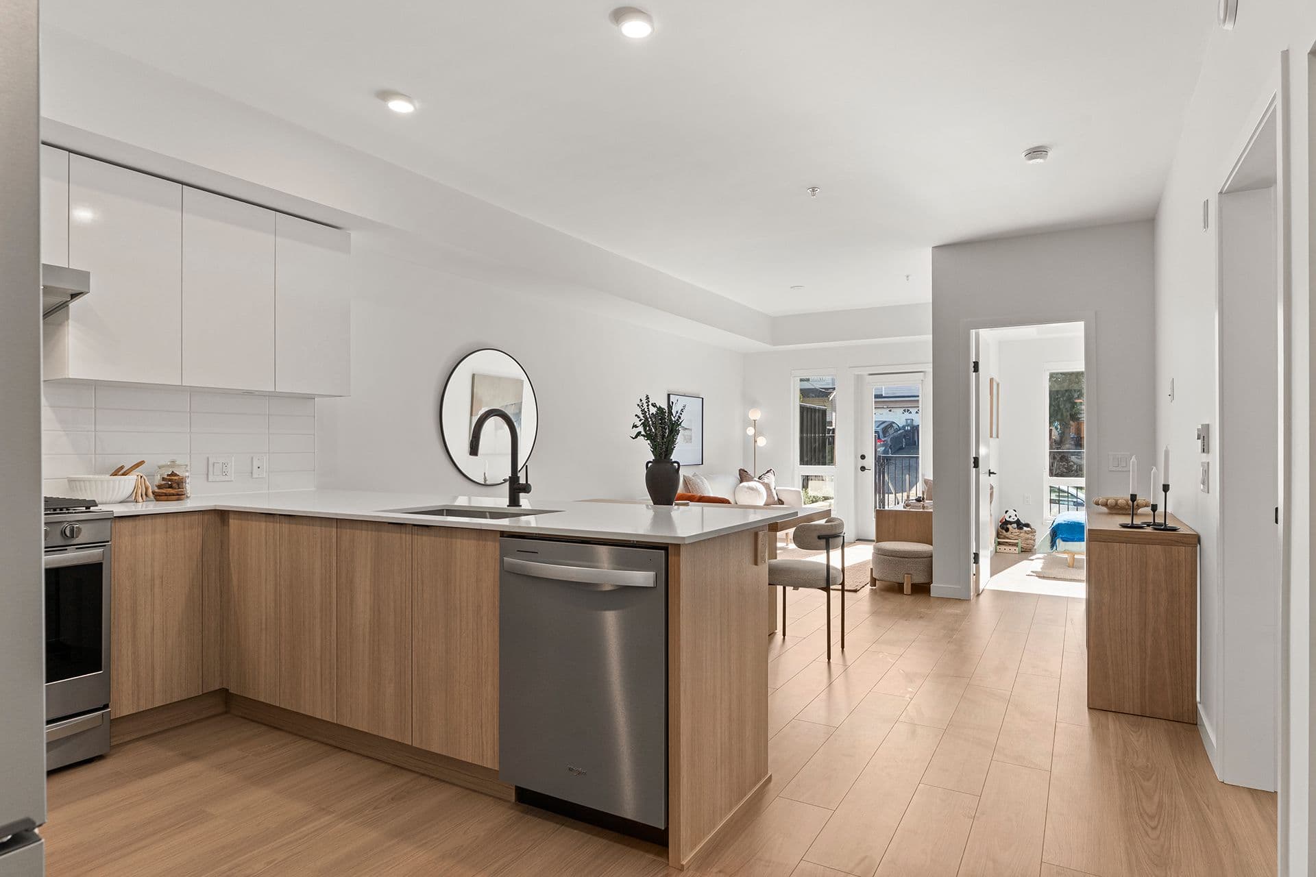 Hawthorne – A bright, open-concept kitchen features light wood lower cabinets, white upper cabinets, white quartz countertops, and stainless steel appliances. The room seamlessly flows into a sunlit living area and an adjoining bedroom in the background, all framed by light wood flooring and minimalist decor.