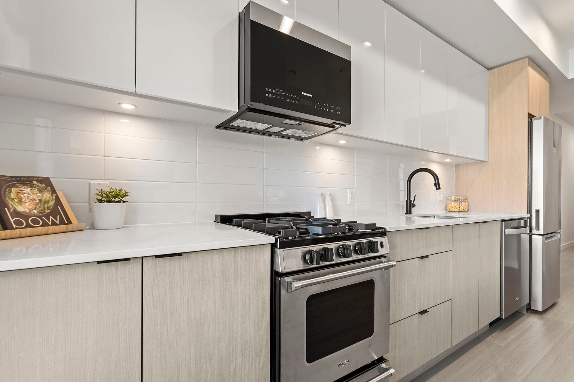Hawthorne – A modern residential kitchen featuring light-wood lower cabinets, glossy white upper cabinets, and a white tile backsplash under warm recessed lighting. Stainless steel appliances include a gas range, over-the-range microwave, integrated dishwasher, and refrigerator, paired with white quartz countertops and a sleek black goose-neck faucet.