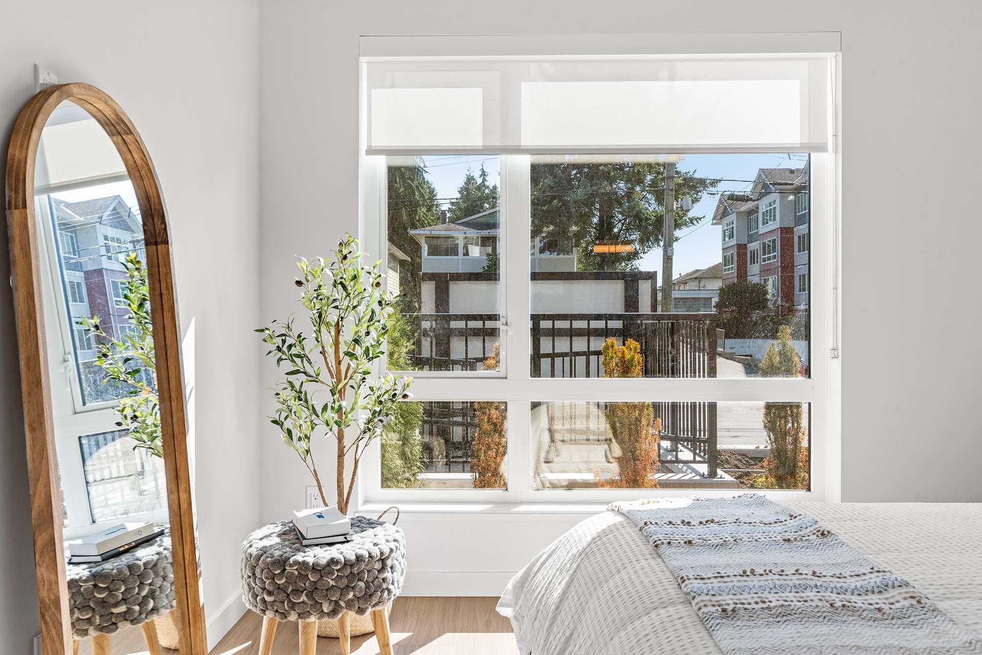 Hawthorne – A bright bedroom interior featuring a large window with white roller shades that overlook neighbouring buildings and trees. The room is styled with a full-length arched wooden mirror, a small potted olive plant, a textured ottoman, and a bed draped with a woven throw blanket.
