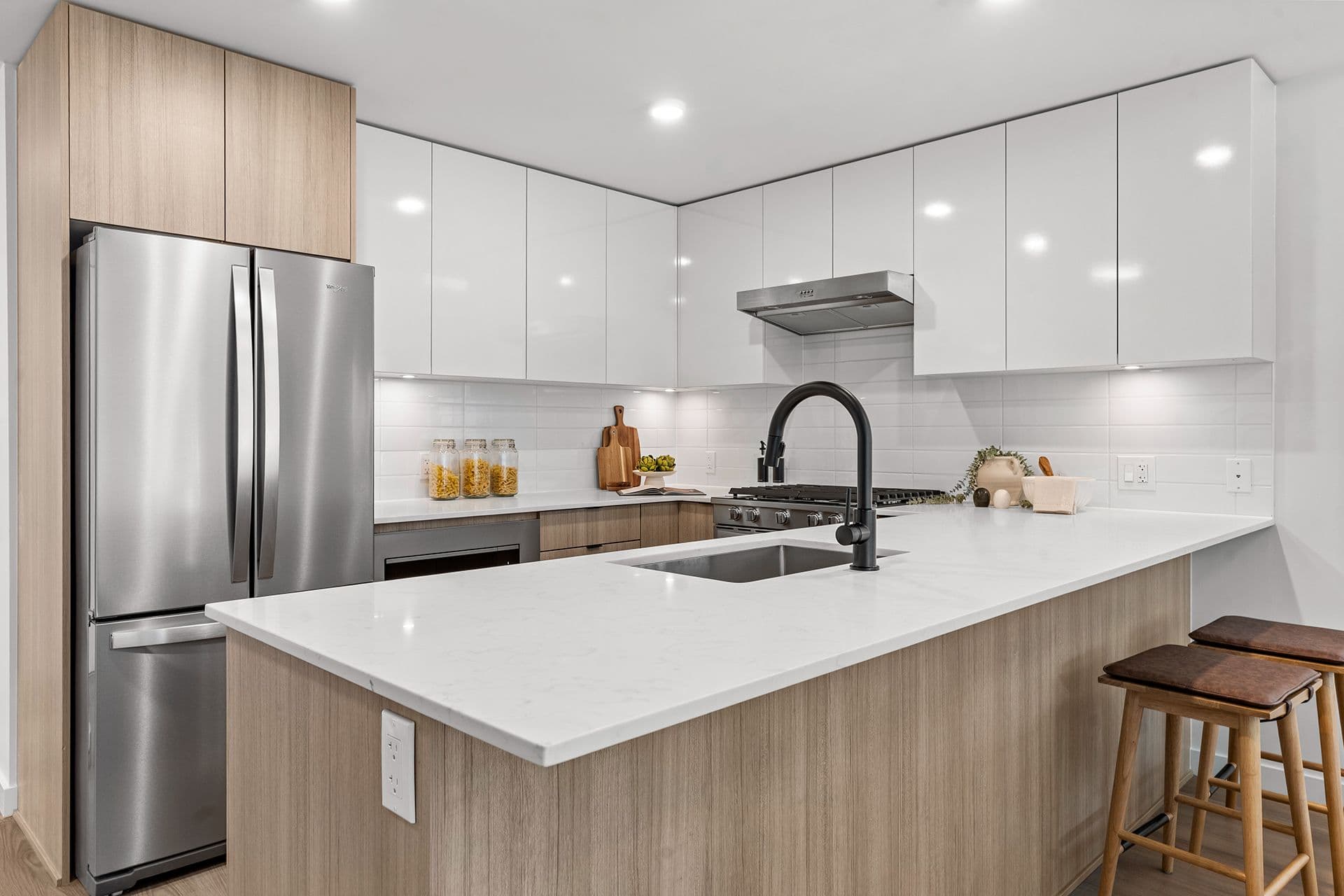 Hawthorne – A modern residential kitchen featuring a large kitchen island with light quartz countertops, a matte black faucet, and light wood cabinetry underneath paired with wooden barstools. Stainless steel appliances include a french-door refrigerator and gas range, complemented by white upper cabinetry and a subway tile backsplash illuminated by under-cabinet lighting.