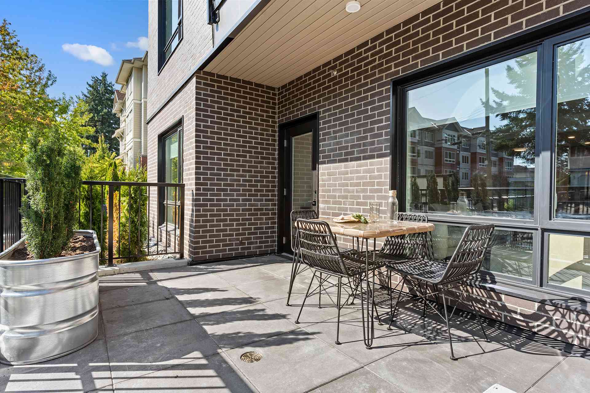 Hawthorne – A ground-floor outdoor patio features dark stone pavers, a small woven glass-top dining table with black chairs, and a corrugated metal planter with lush green shrubs against a dark brick building facade. Bright daylight casts crisp shadows across the space, with neighboring residential buildings and mature trees visible in the background.