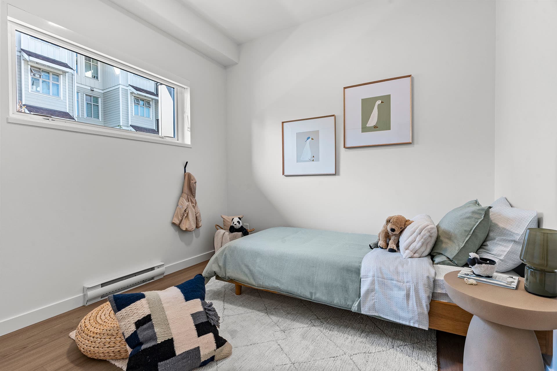 Hawthorne – A modern child's bedroom featuring a single wooden bed dressed in sage green and light neutral linens, set over light wood flooring and a textured area rug. White walls are accented with framed bird artwork, a high horizontal window letting in natural light, and playful decor like stuffed animals and a woven pouf.