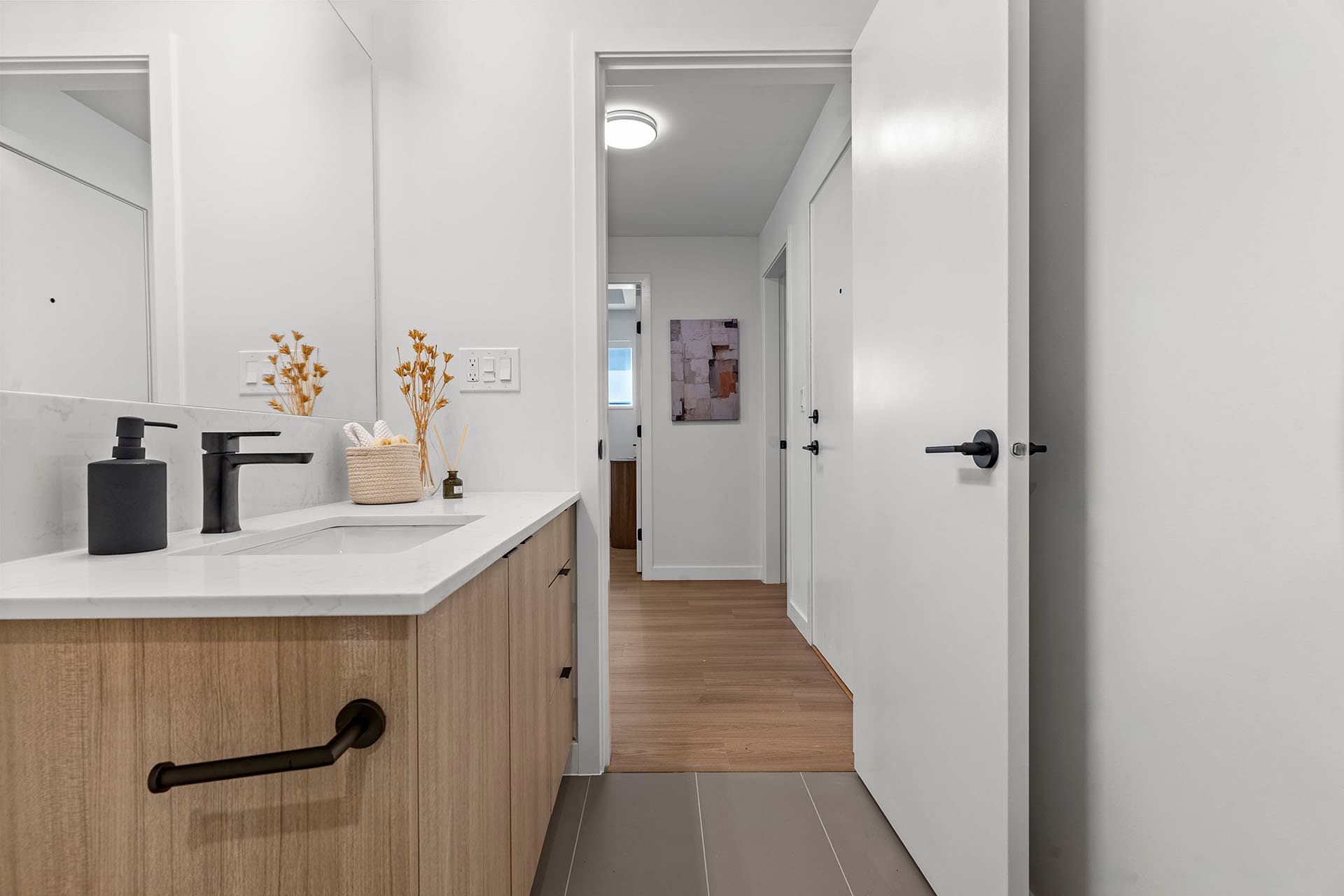Hawthorne – A modern residential bathroom vanity with light wood-tone cabinetry, a white quartz countertop, matte black hardware, and gray floor tiles. The open bathroom doorway offers a view into a hallway with light hardwood floors and bright white walls.