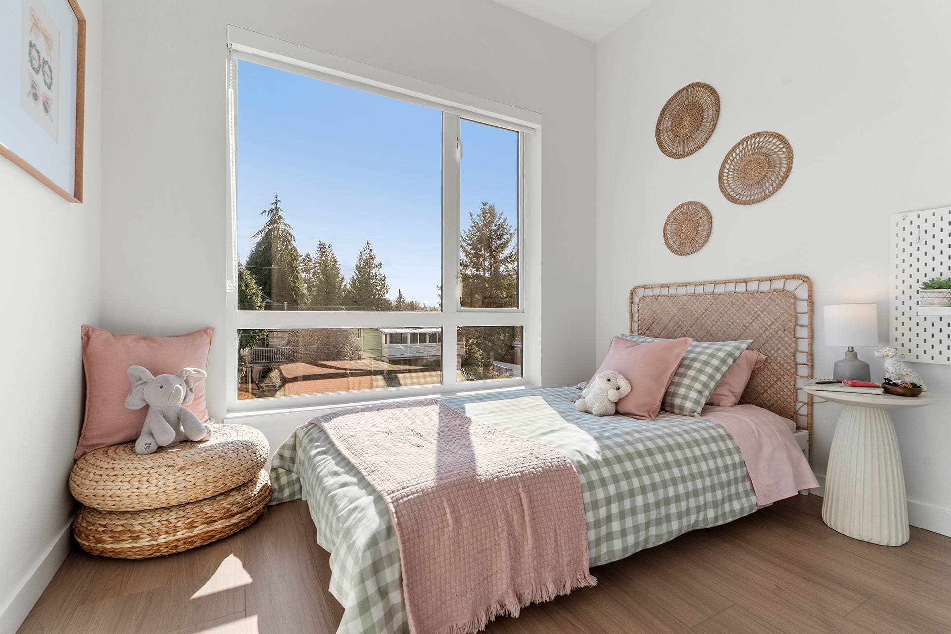 Hawthorne – A bright, sunlit child's bedroom features a bed dressed in green gingham and soft pink bedding set against a rattan headboard. A large window brings in daylight and looks out onto treetops, while natural woven accents like wall baskets and stacked floor cushions complete the warm, cozy space.