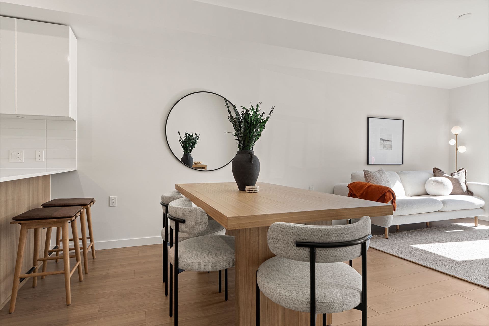 Hawthorne – An open-concept living and dining area featuring light wood flooring, a wooden dining table with upholstered modern chairs, and a kitchen peninsula with barstools. White walls, soft natural light, a large circular mirror, and a bright living room setup with a beige sofa create a clean, contemporary aesthetic.