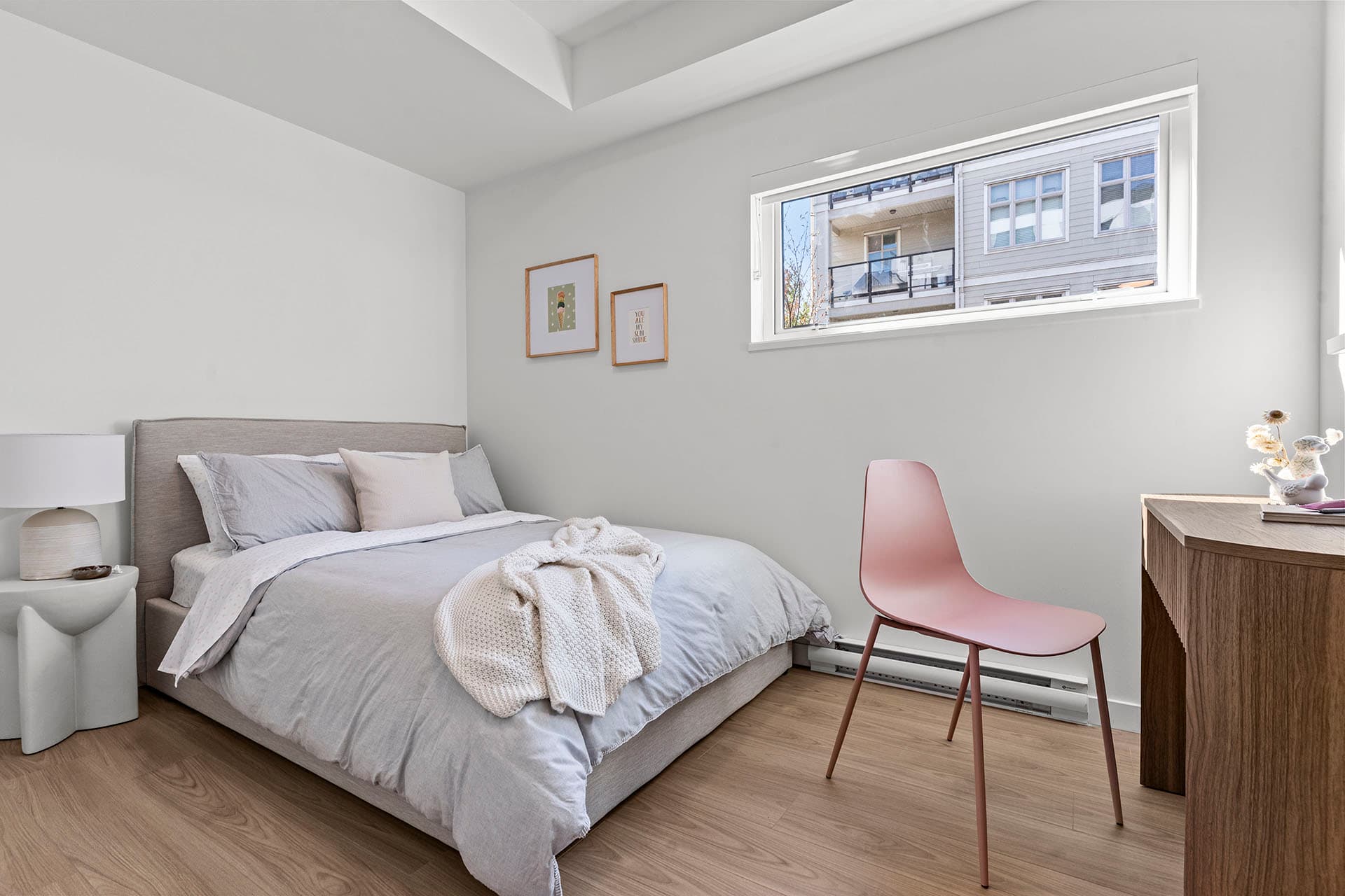 Hawthorne – A modern, minimalist bedroom features a bed made up with light gray and blush bedding alongside a wooden desk paired with a dusty pink chair. Light oak laminate flooring and white walls create a bright, airy atmosphere, complemented by natural light coming through a high horizontal window.