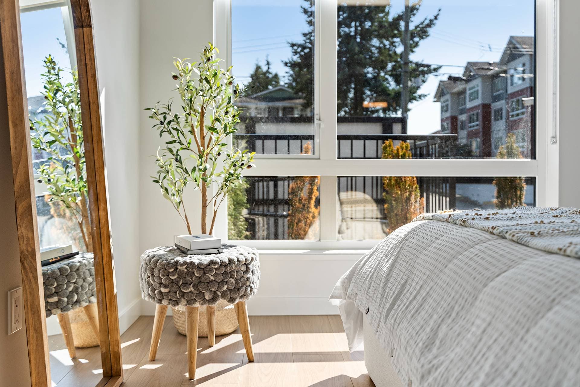 Hawthorne – A sunlit bedroom corner features a wooden full-length standing mirror, a small potted olive tree, and a grey felt-pompom stool set against light wood flooring. Large windows flood the space with natural daylight and offer a view of neighbouring residential buildings and trees outside.
