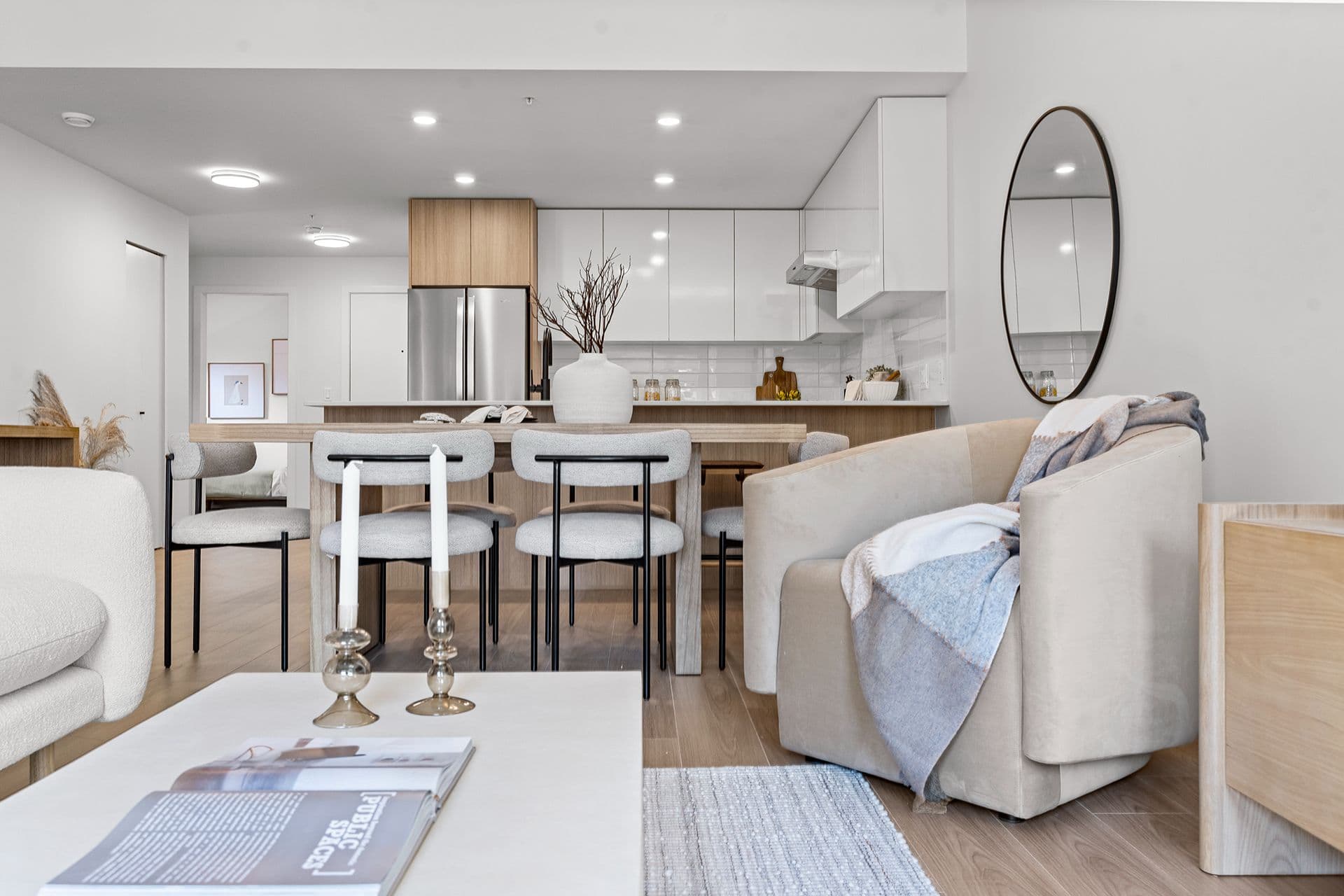 Hawthorne – A modern open-concept interior view stretching from a cozy living area with a plush beige armchair in the foreground to a wooden dining table and an open kitchen with white upper cabinets and stainless steel appliances. The space is styled in bright neutral tones with light wood finishes, polished white surfaces, and subtle decorative accents.