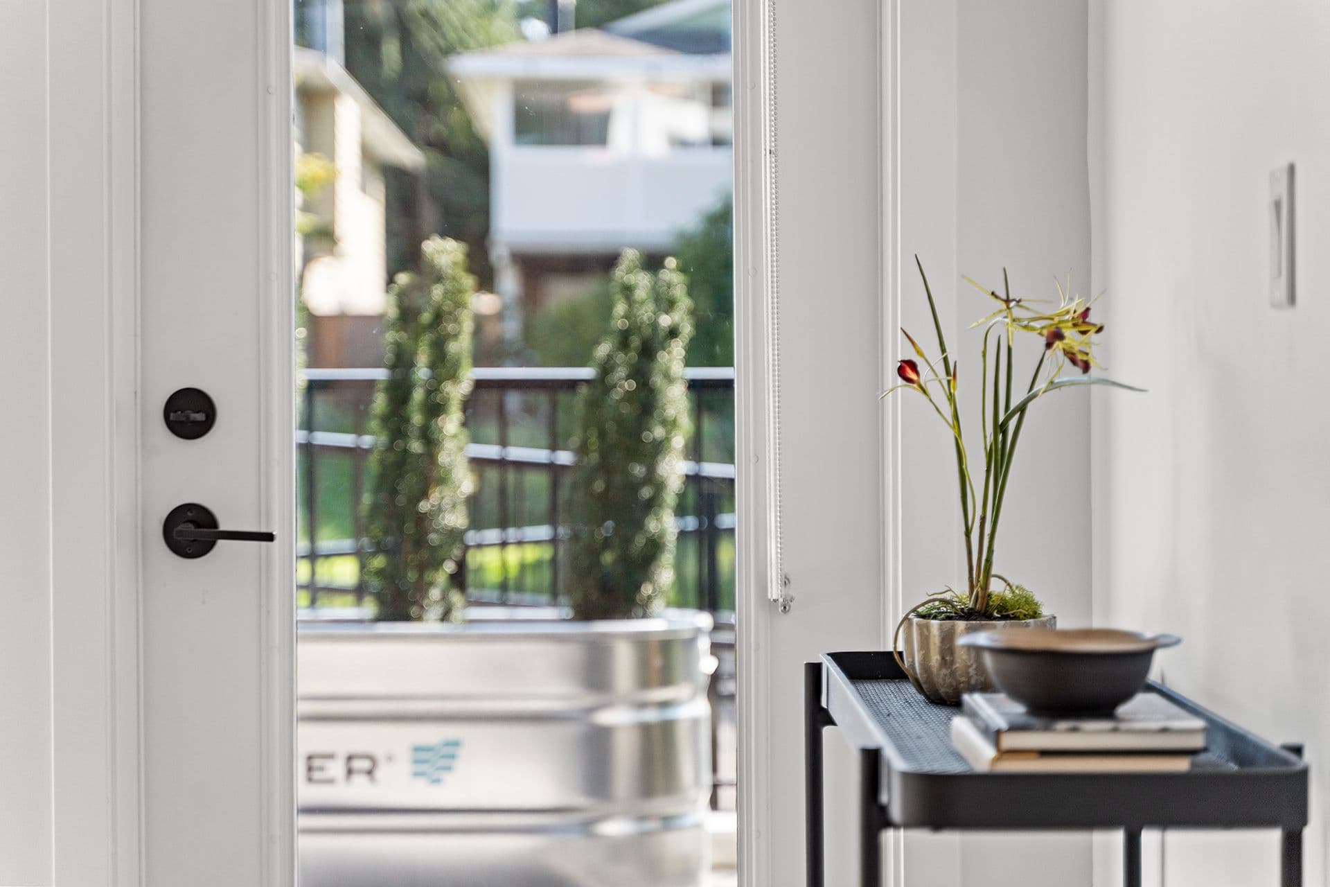 Hawthorne – An open white door frame with matte black hardware offers a view onto a private patio featuring a large metal trough planter with small evergreen trees. In the foreground interior, a delicate potted orchid and a stack of books sit on a sleek black side table against bright white walls.