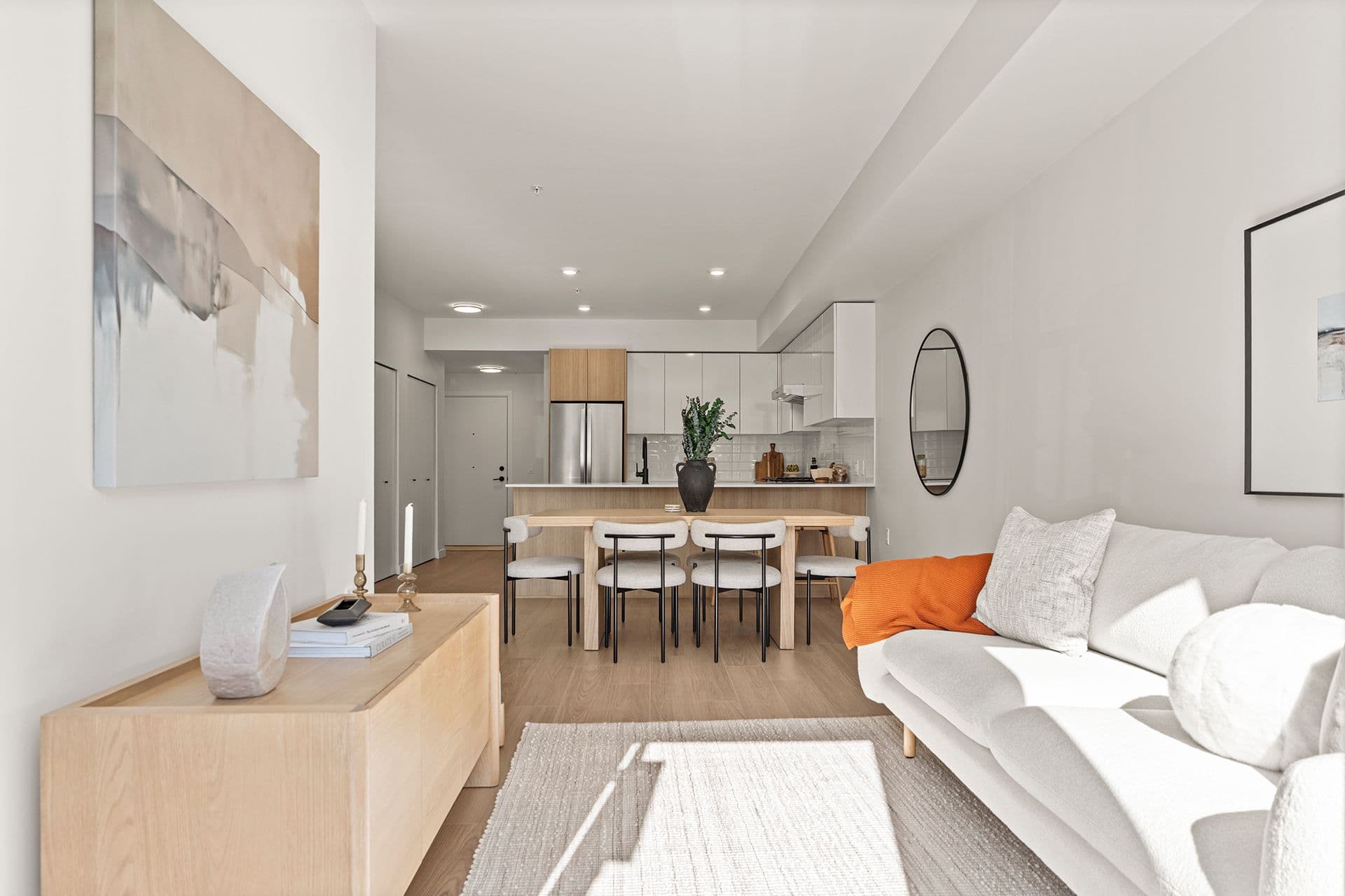 Hawthorne – A brightly lit, open-concept living and dining area leads into a modern kitchen with light wood accents and white cabinetry. The space is styled in neutral tones featuring a cream sofa, a wooden dining table with six chairs, light oak flooring, and subtle pops of color like an orange throw blanket.