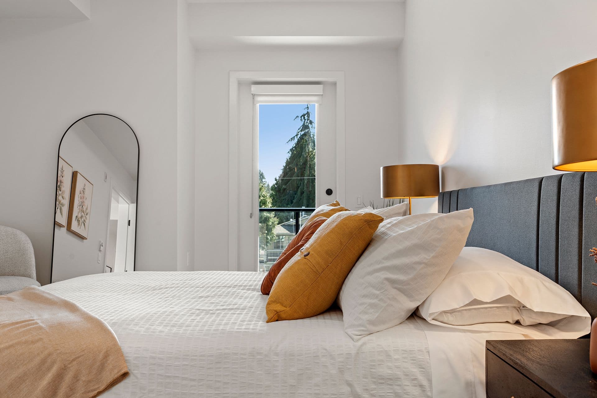 Hawthorne – A modern, sunlit bedroom featuring a neatly made bed with white waffle-weave bedding, warm yellow and orange accent pillows, and a grey upholstered headboard flanked by golden bedside lamps. Plain white walls frame an arched full-length floor mirror and a narrow doorway leading out to a balcony overlooking lush green trees.