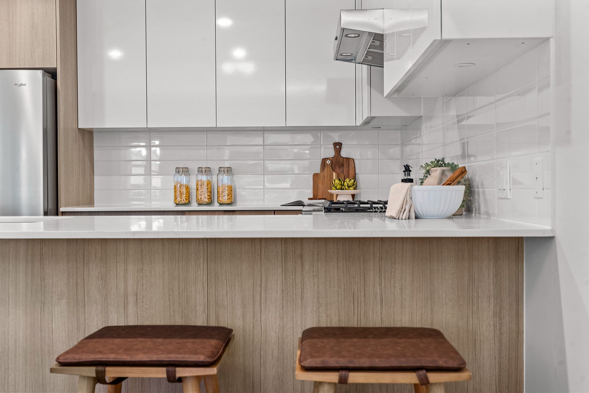 Hawthorne – A modern condo kitchen featuring glossy white upper cabinets, light wood grain lower paneling, and two brown leather barstools at the quartz island. The countertop is neatly styled with glass storage jars, wooden cutting boards, and decorative kitchenware against a white subway tile backsplash.