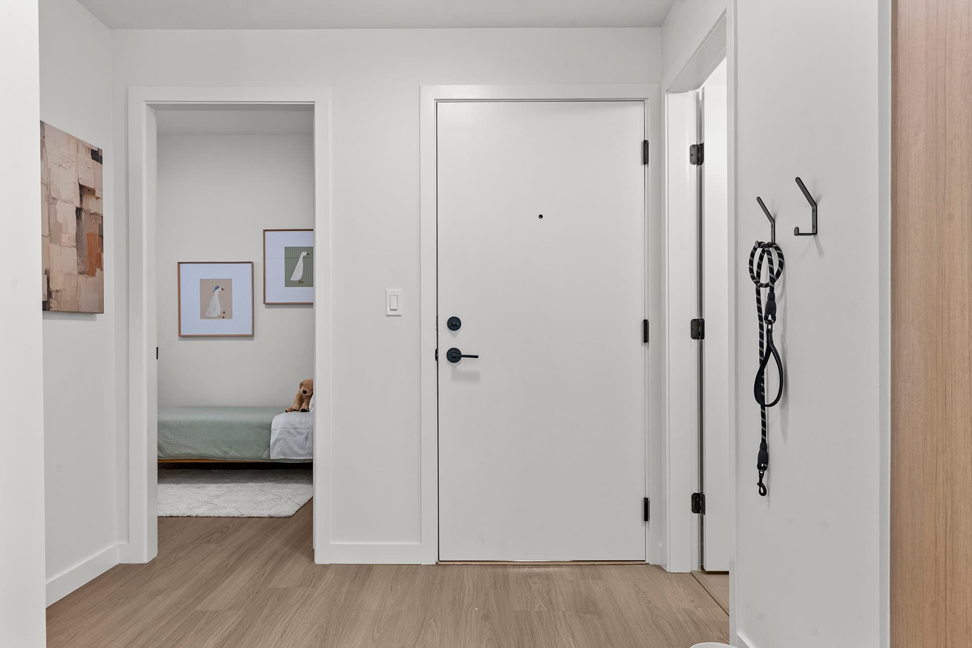 Hawthorne – A clean, modern entryway featuring light wood-look laminate flooring, crisp white walls, and a white front door with matte black hardware. To the left, an open doorway reveals a brightly lit bedroom with framed artwork and a bed, while black wall hooks hold a dog leash near the entrance.